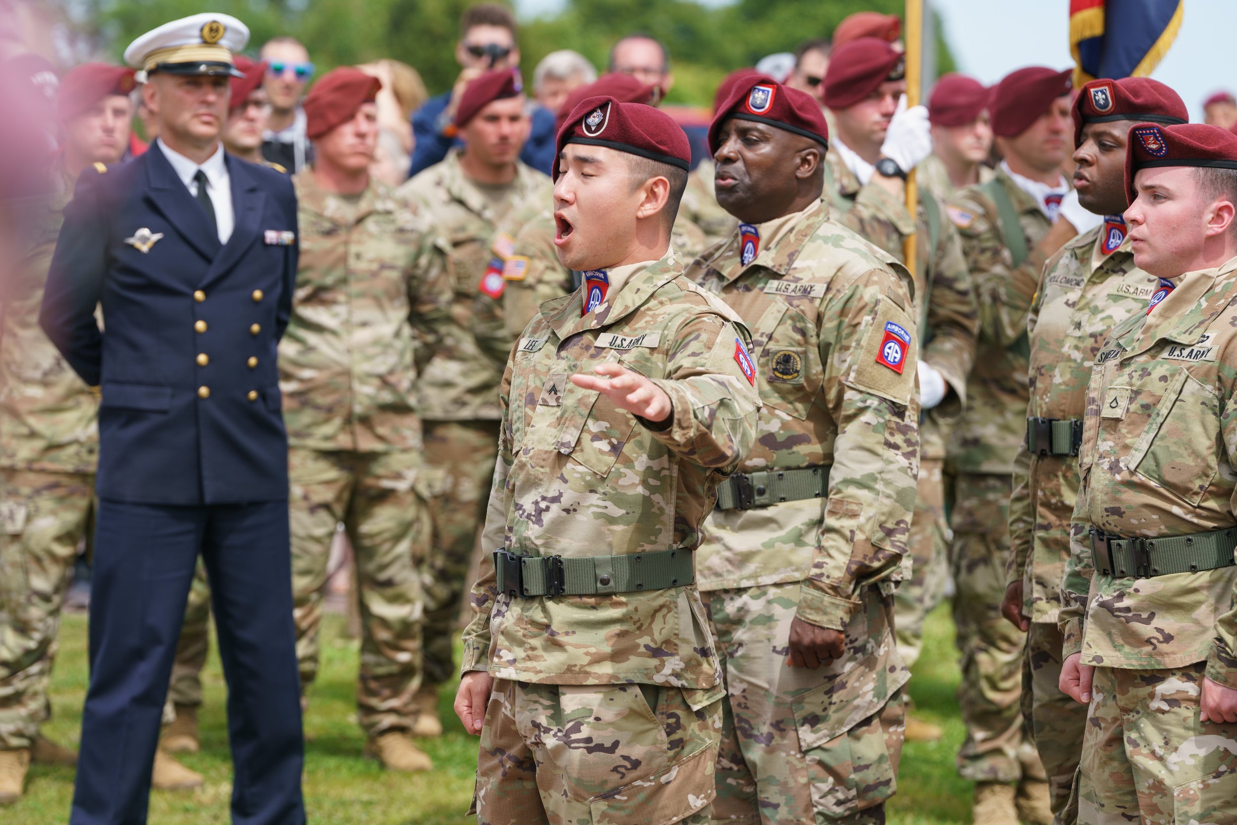 A group of soldiers in camouflage uniforms and red berets are standing at attention during a military ceremony, with a woman in a navy blue uniform observing.
