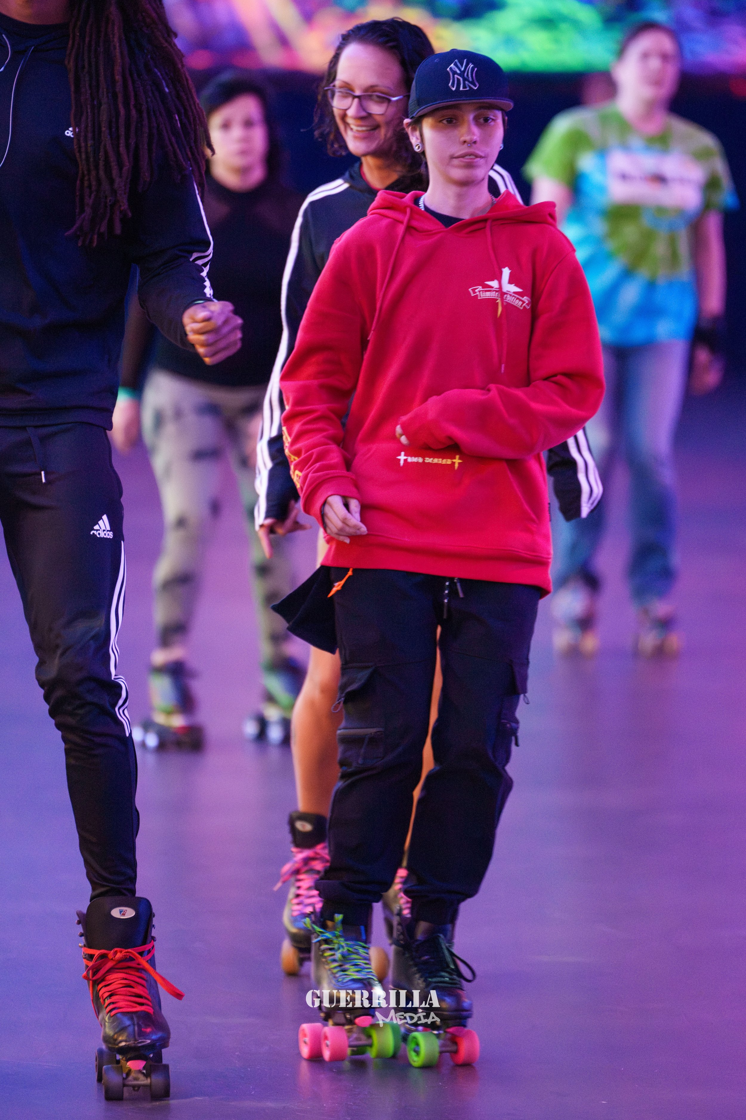 A group of people roller skating indoors, with a woman smiling and wearing glasses, and a young person wearing a red hoodie and a black NYC cap in front.