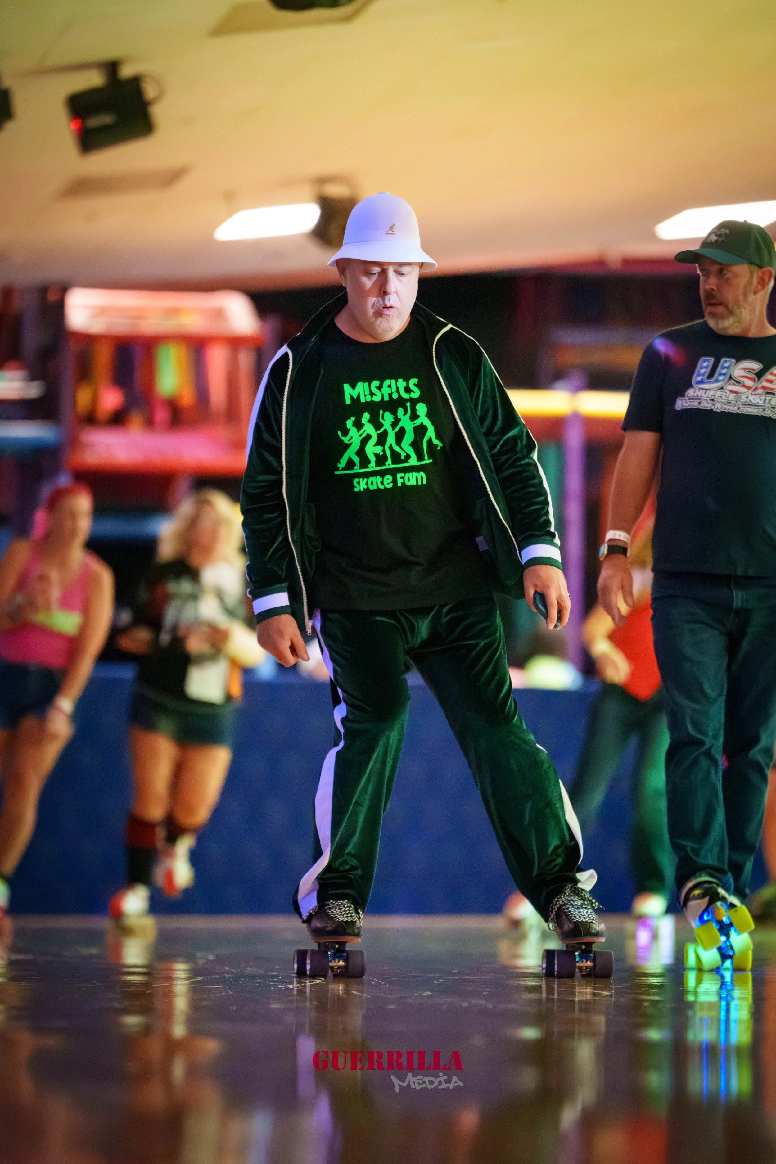 Person roller skating indoors, wearing a black hoodie with a neon green graphic and text, white bucket hat, and black pants.