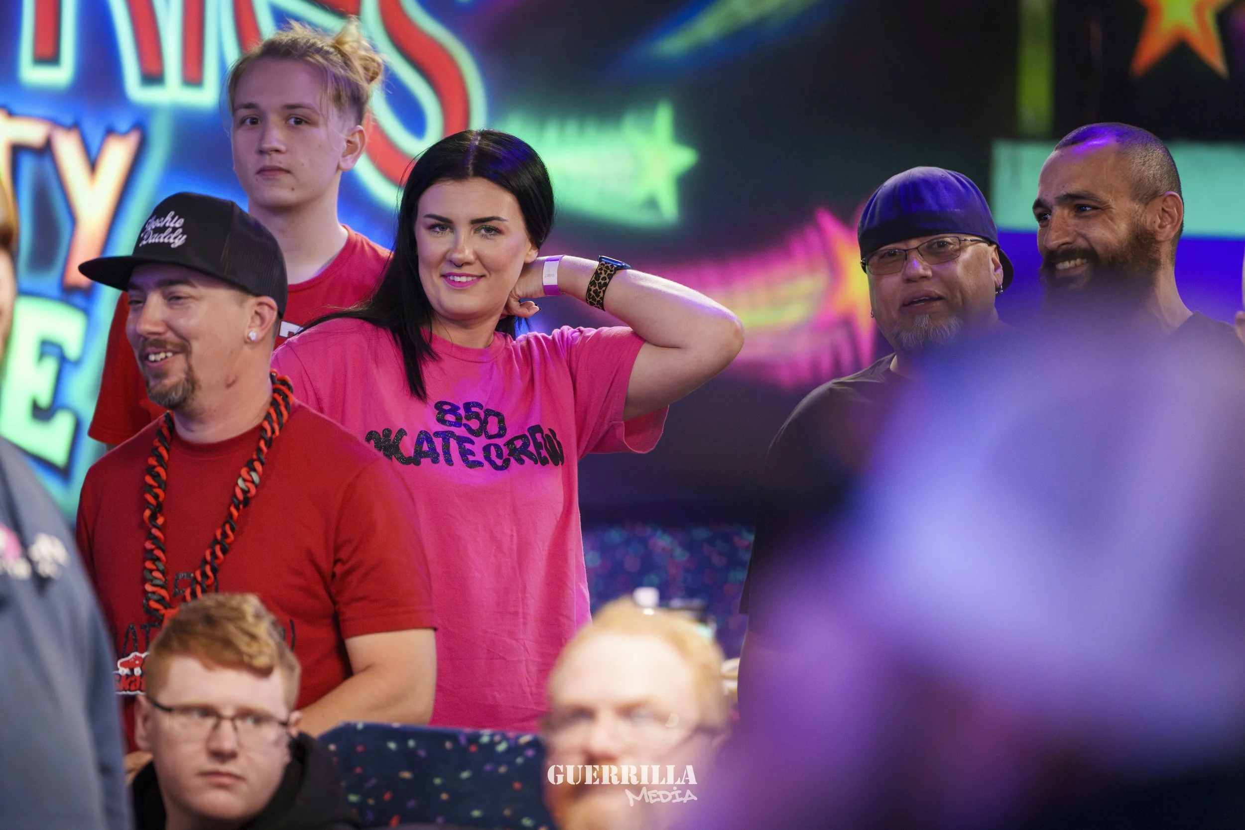 Group of people at some event, with neon lights in the background, participating or watching. The woman in the pink shirt is smiling and adjusting her hair, while other individuals are engaged around her.