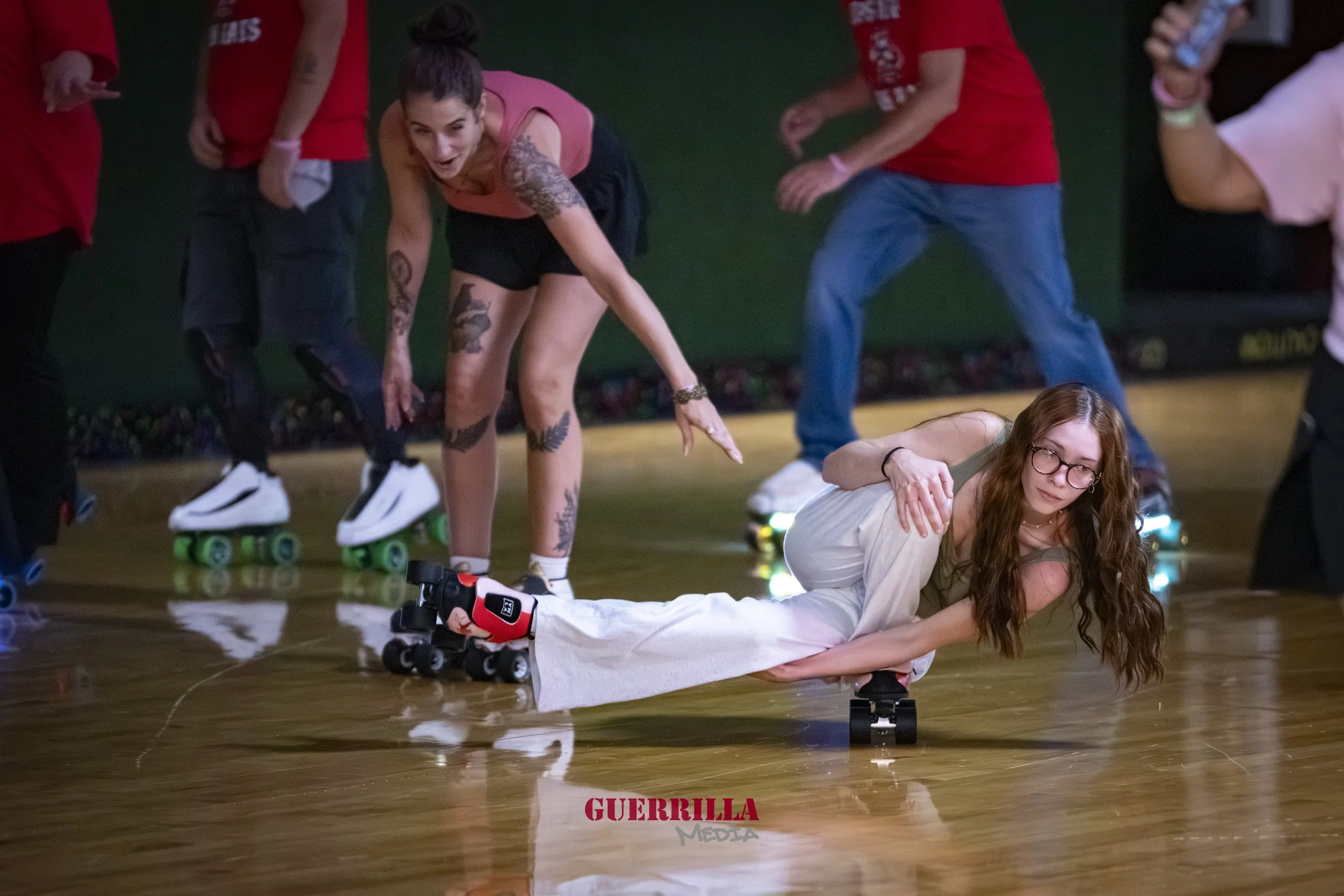 A woman in glasses lying on roller skates on the floor while holding onto the legs of a woman with tattoos, also on roller skates, who is leaning forward. Other people with roller skates are visible in the background, some holding water bottles.