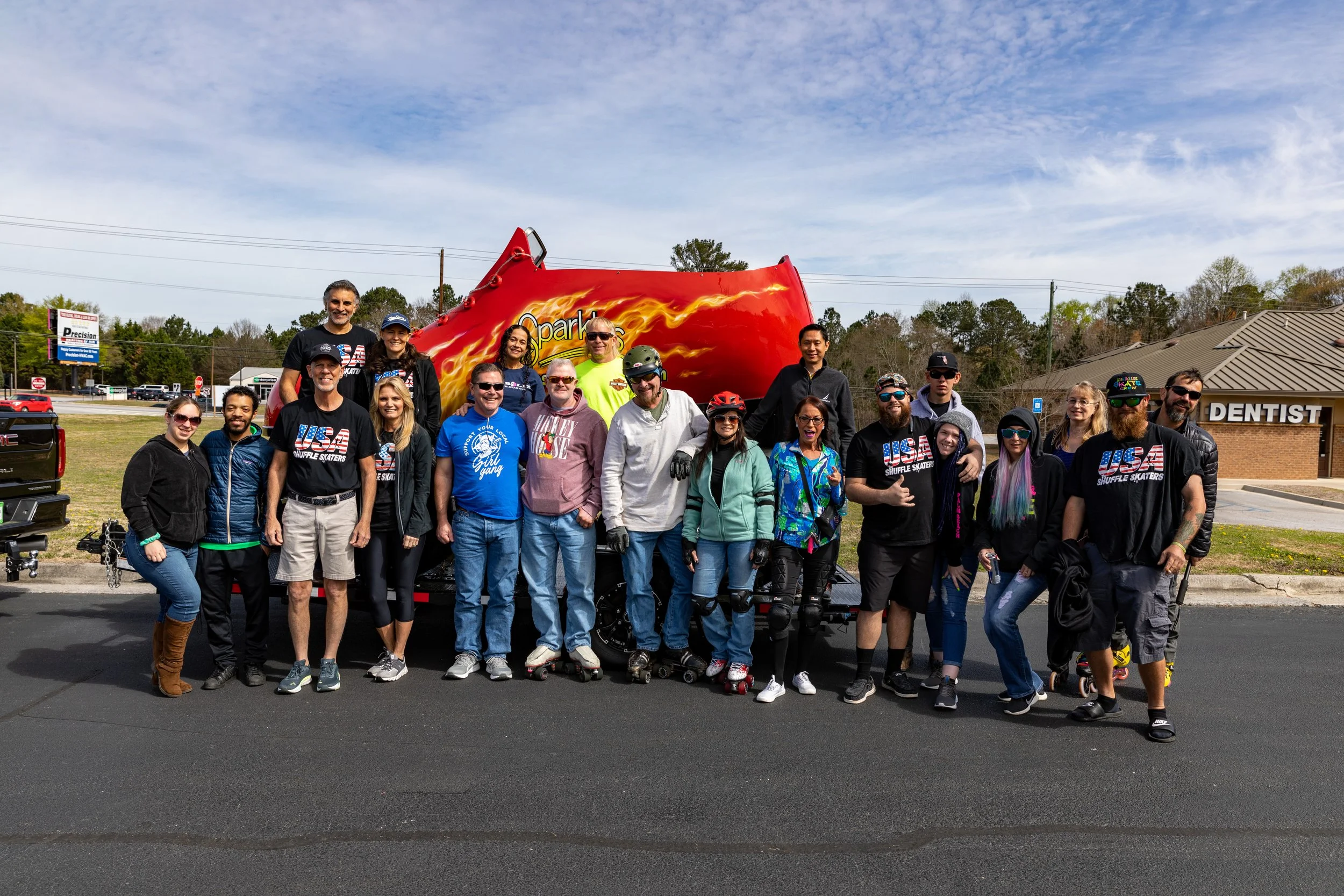 Group of people posing in front of a large red graphic shoe with flames, on a street near a dentist office, with a building and trees in the background.