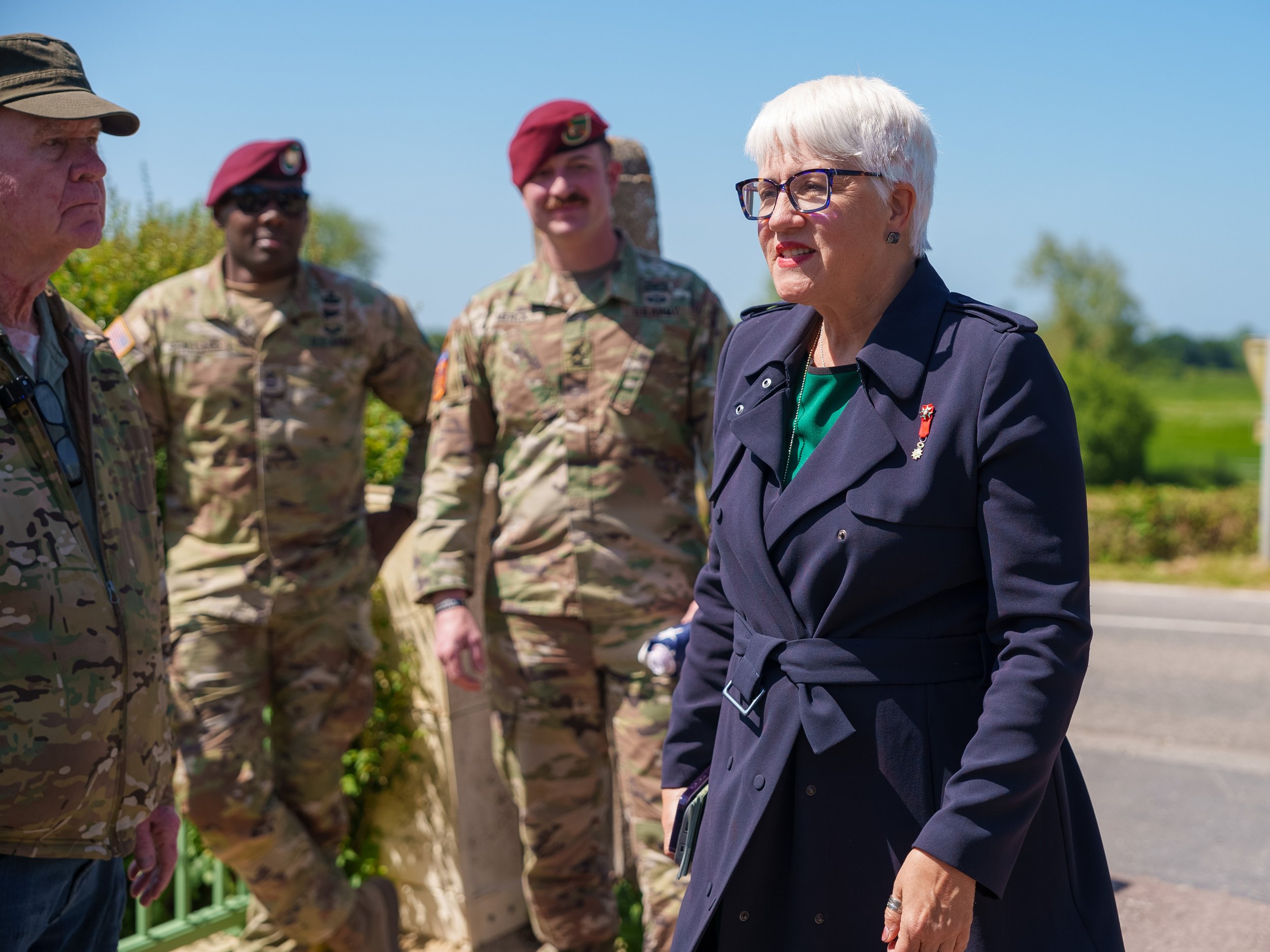 A woman in a navy trench coat and glasses engaging in conversation with military personnel outdoors on a sunny day.