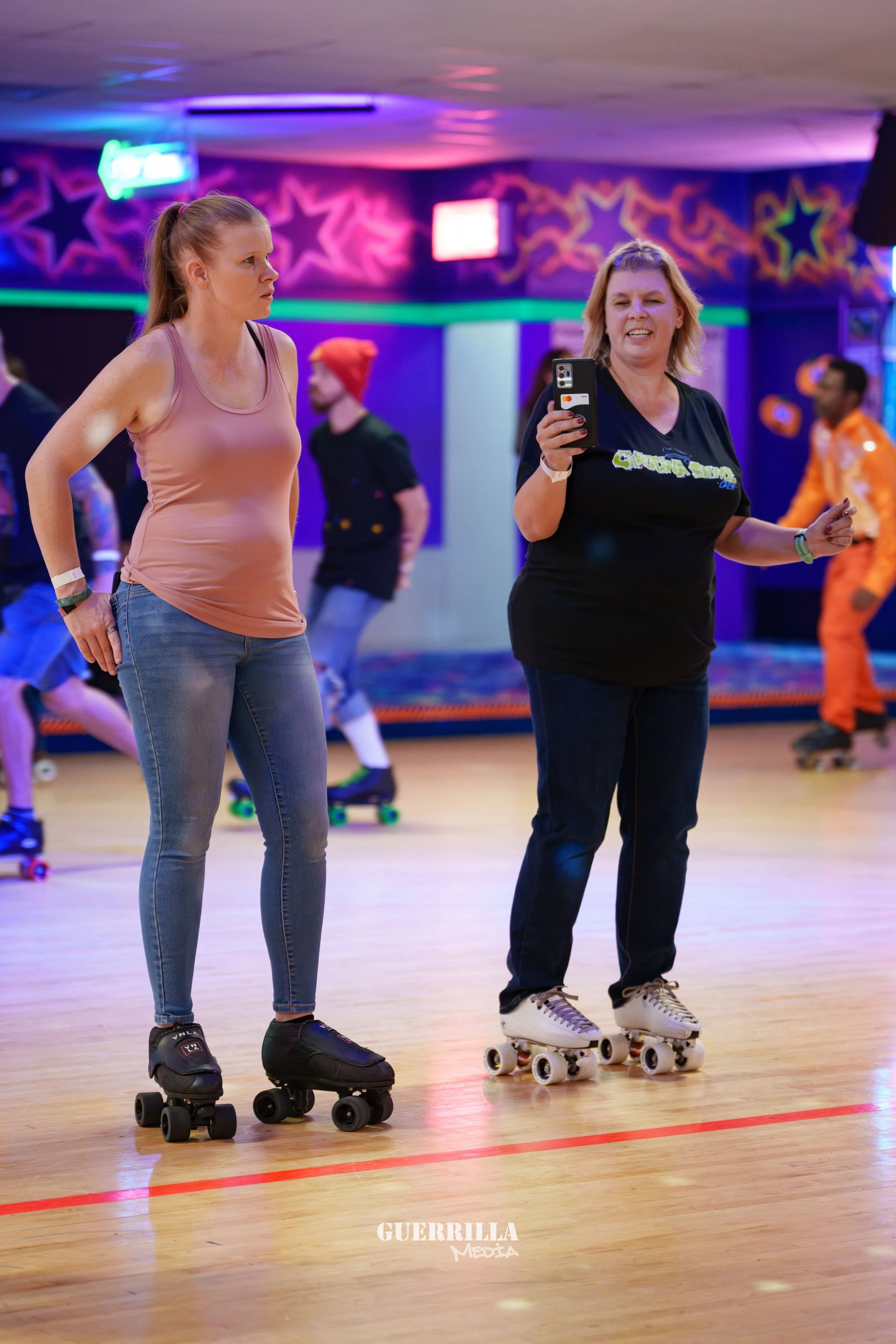 Two women roller skating in an indoor roller skating rink with colorful neon lighting and star decorations on the wall. One woman is wearing a black shirt and jeans, holding a phone, smiling, while the other woman in a pink tank top and jeans looks o