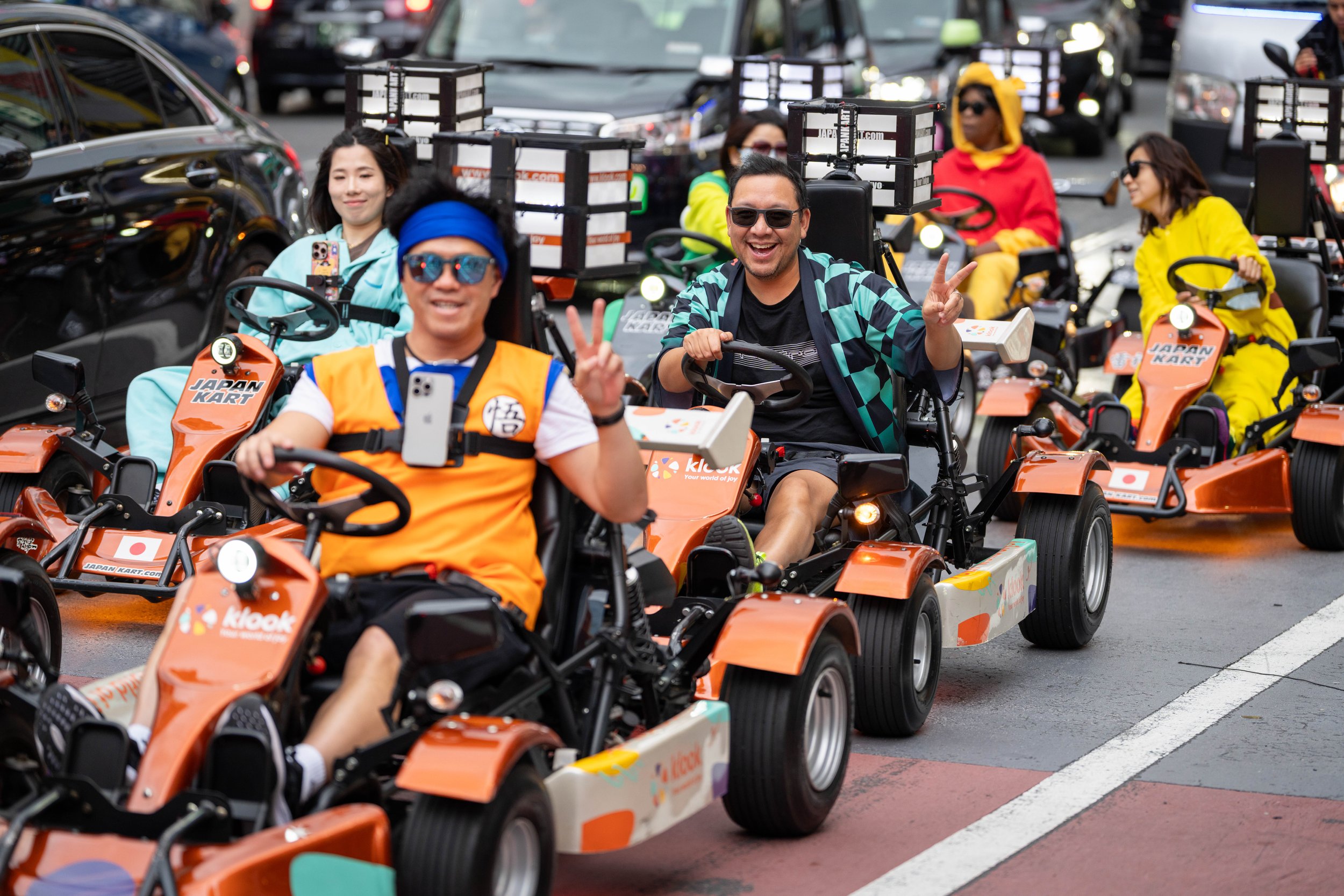 People riding orange golf cart-style vehicles labeled "Japan Kary" during a parade or event, with smiling and waving participants.