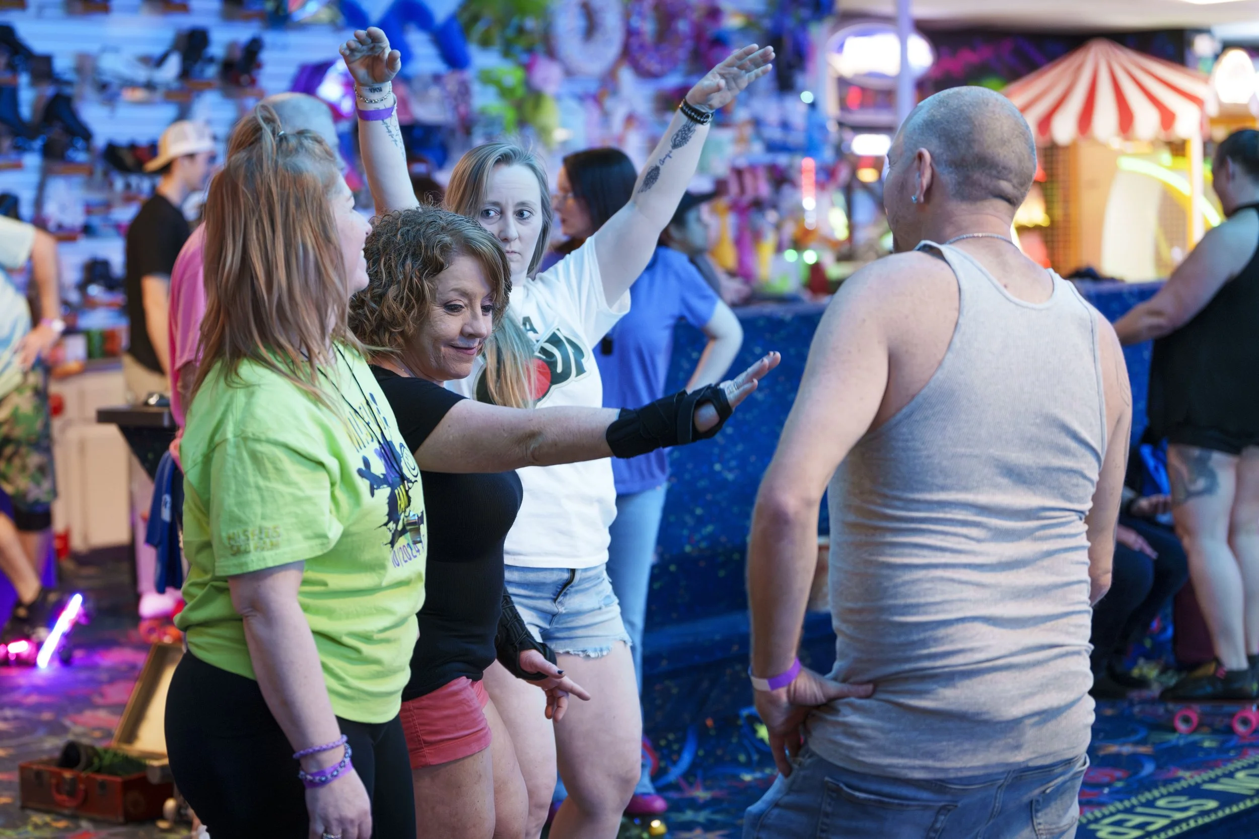 People having fun at an indoor amusement park or arcade, with a man in a gray tank top talking to four women, one of whom is reaching out to him.