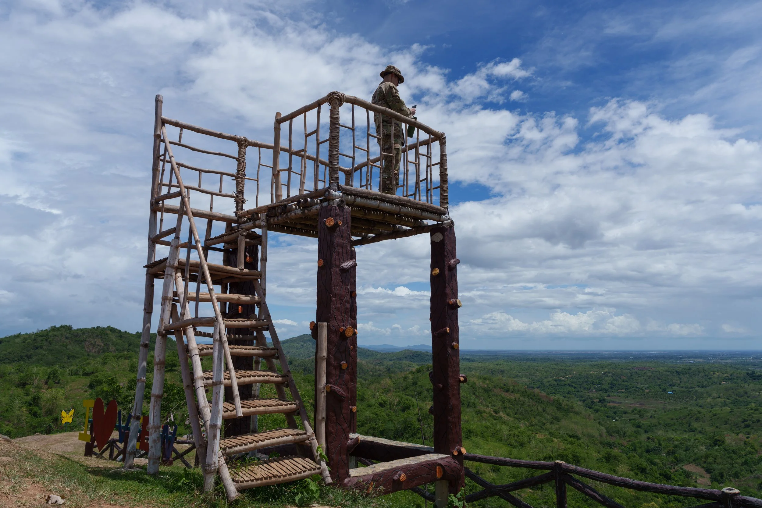 A person in camouflage clothing and a hat standing on a wooden observation tower while looking at a smartphone, overlooking a green landscape with rolling hills and a cloudy sky.