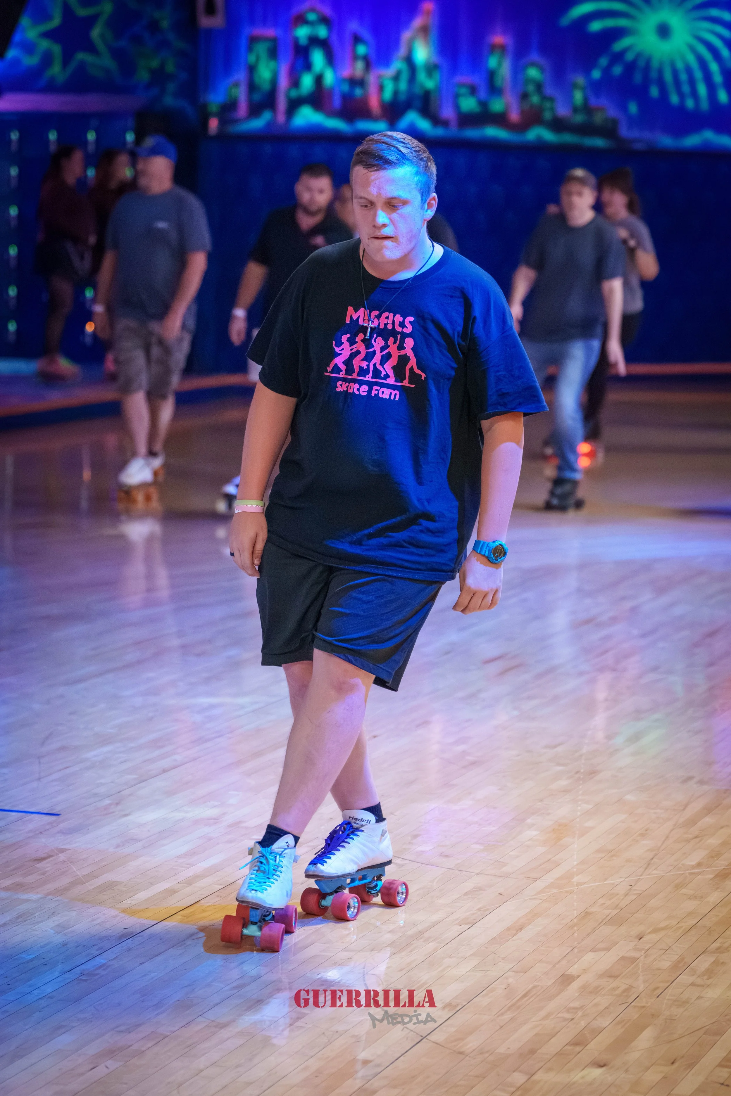 A young man roller skating on a wooden rink with neon lights and a cityscape mural in the background, wearing a black t-shirt and shorts.