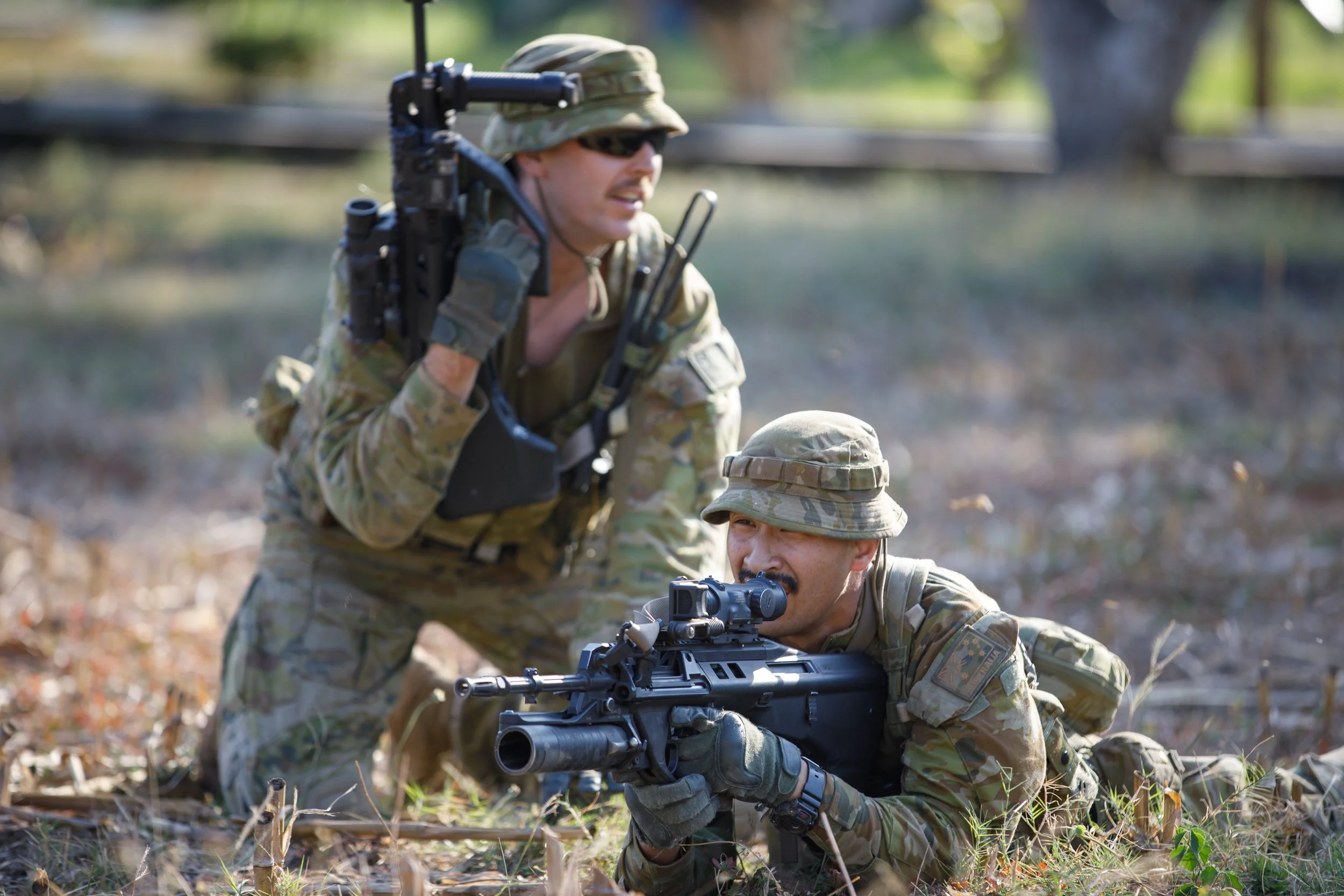 Two soldiers in camouflage uniforms conducting a training exercise outdoors, one lying prone with a sniper rifle and the other kneeling with a radio, in a forested area.