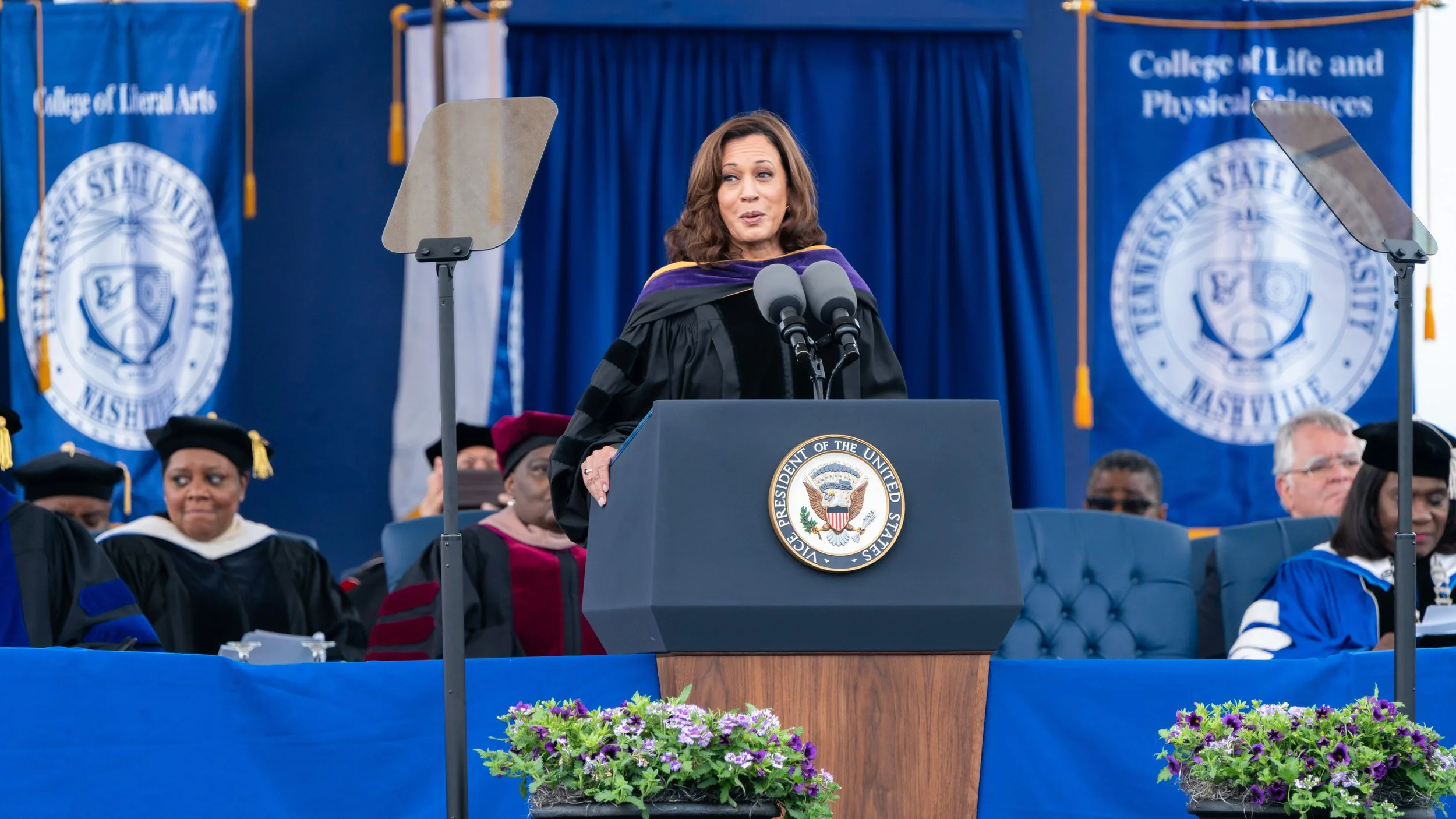A woman in academic regalia speaking at a podium with the presidential seal, at a graduation ceremony at the College of Liberal Arts and College of Life and Physical Sciences, with university banners in the background and seated graduates in academic