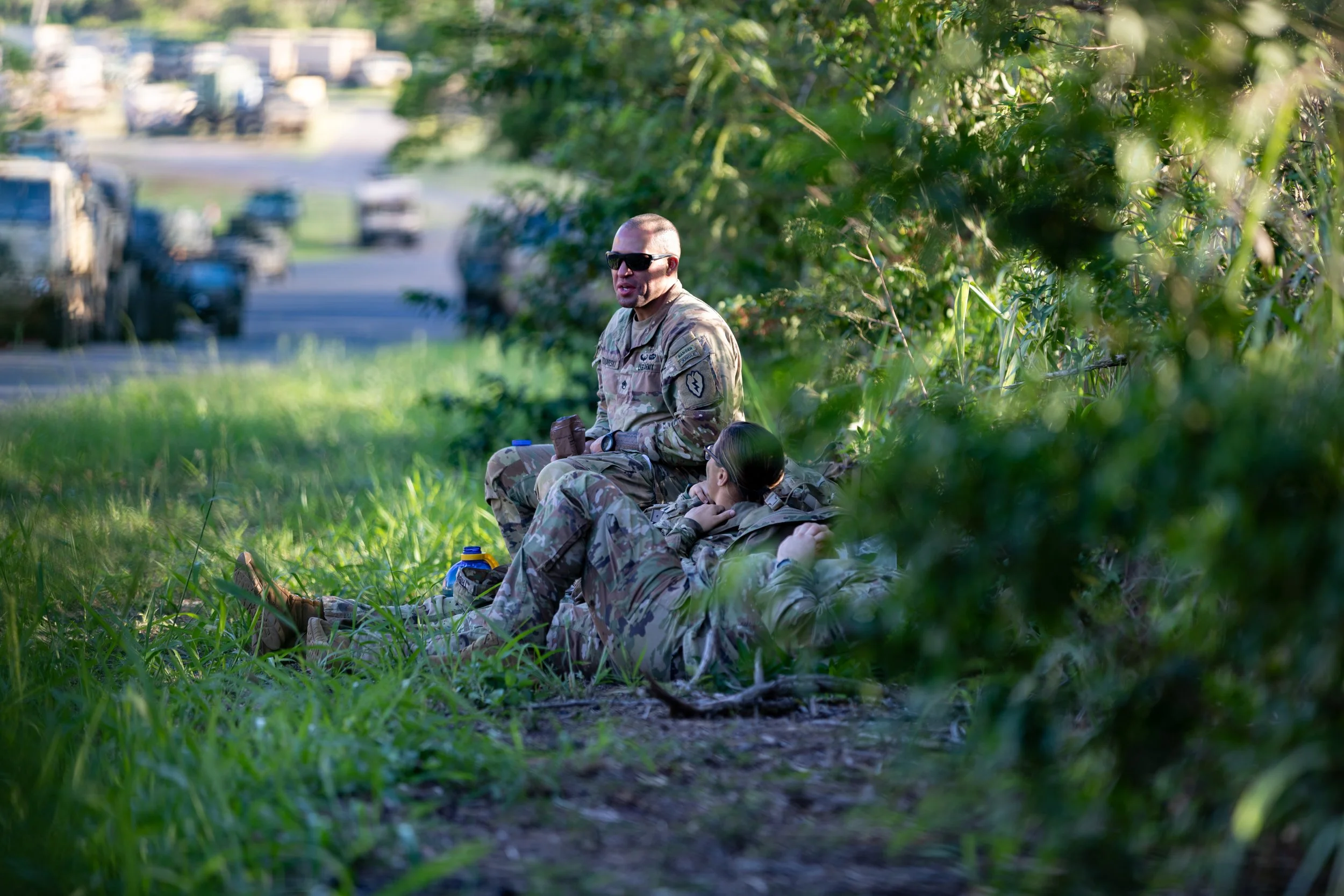 Two soldiers in camouflage uniforms resting in a grassy area behind bushes, with a man wearing sunglasses sitting upright and a woman lying down, smoking a cigarette; vehicles in the background.