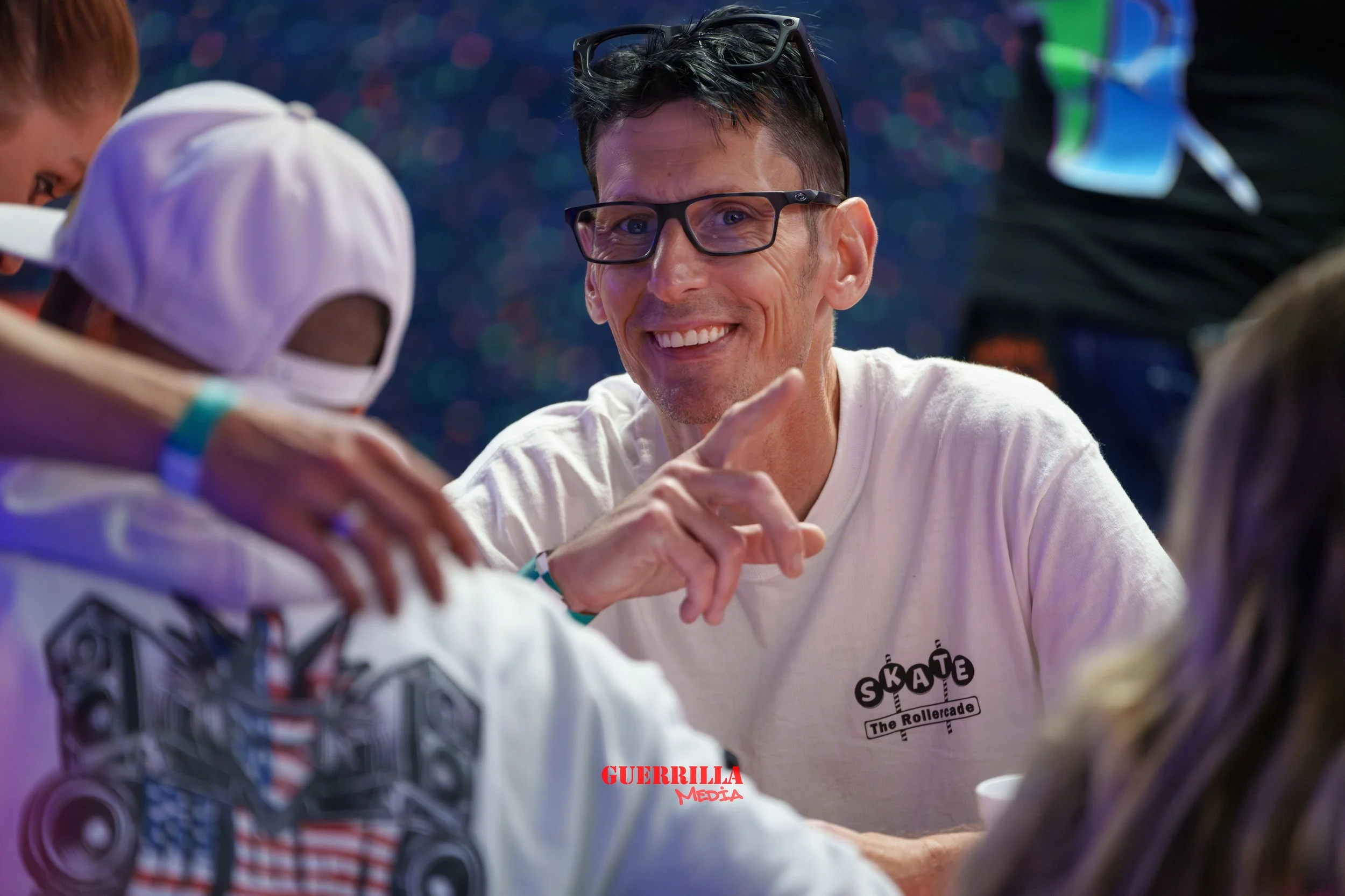 A man with glasses smiling and pointing, sitting with three children at a table, with a colorful, bokeh background, and the text 'GUERRILLA MEDIA' in red at the bottom.