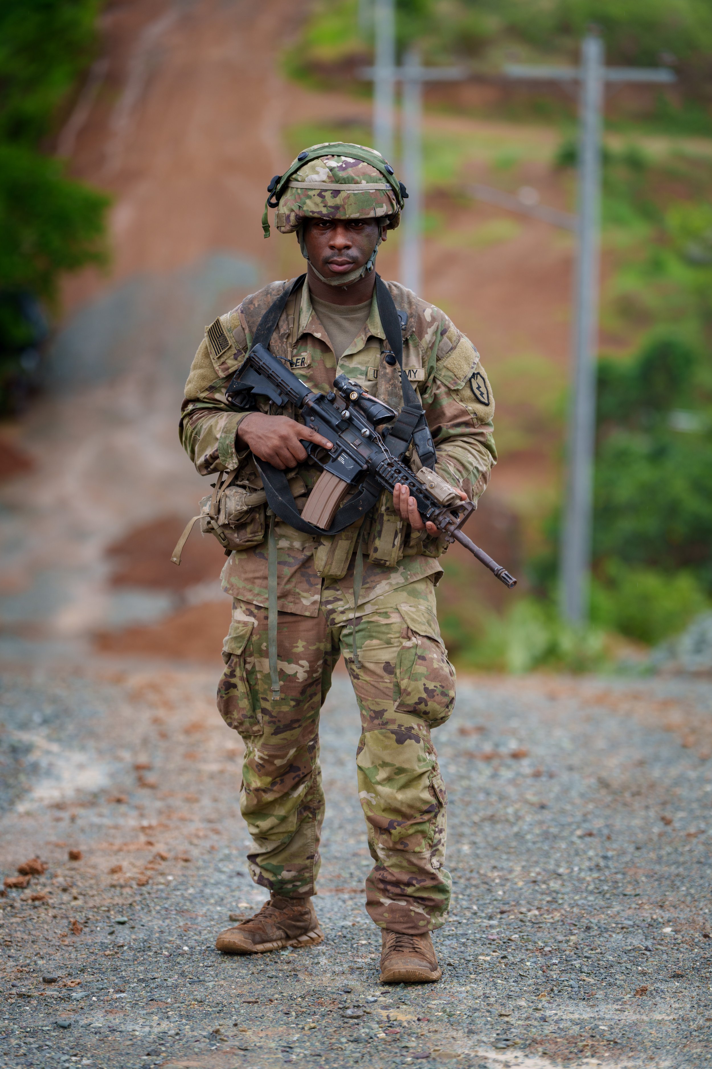 A soldier in camouflage uniform holding a rifle, walking on a dirt road with greenery in the background.