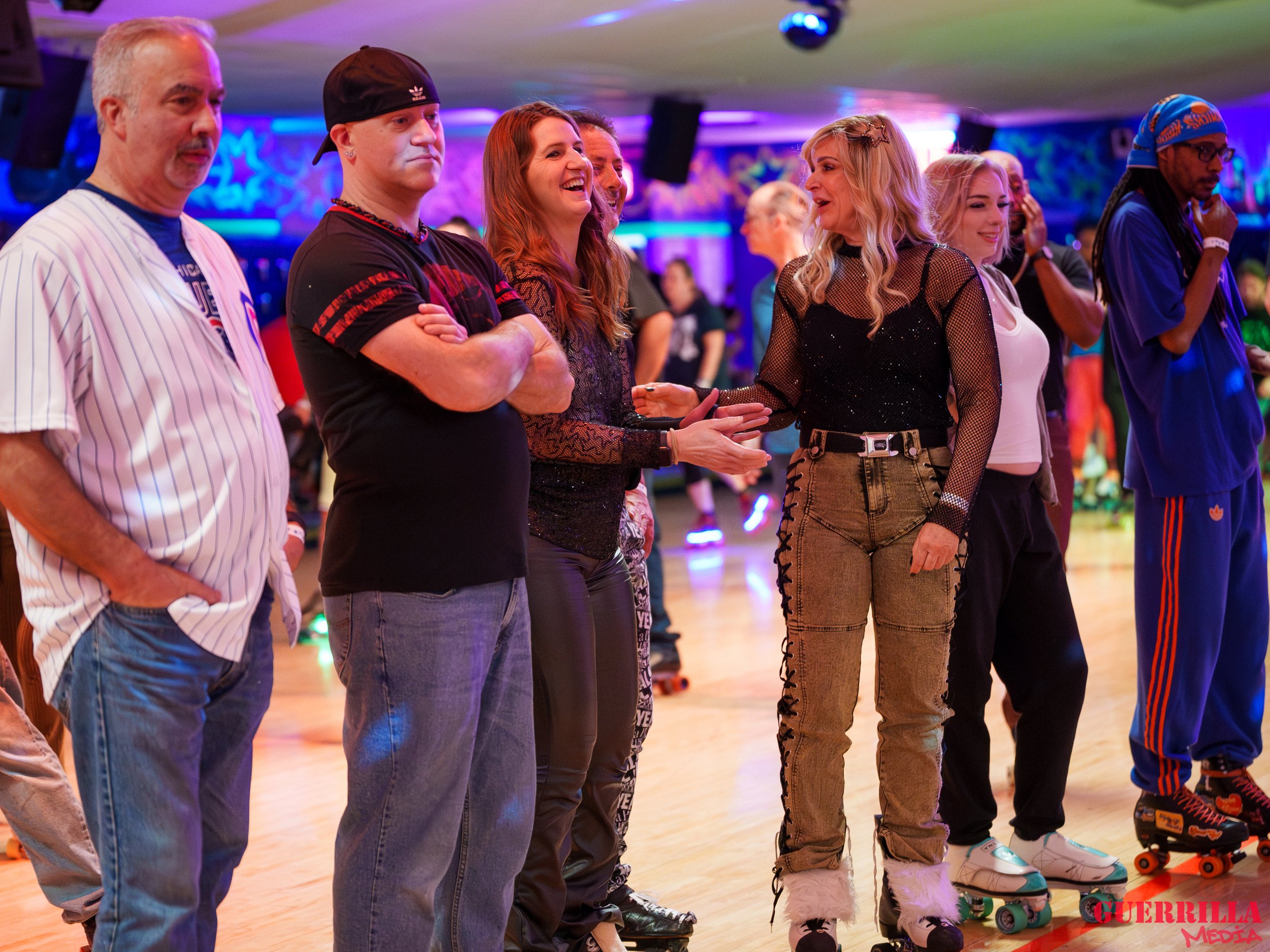 Group of people roller skating indoors, some are chatting and smiling, with colorful neon lights in the background.