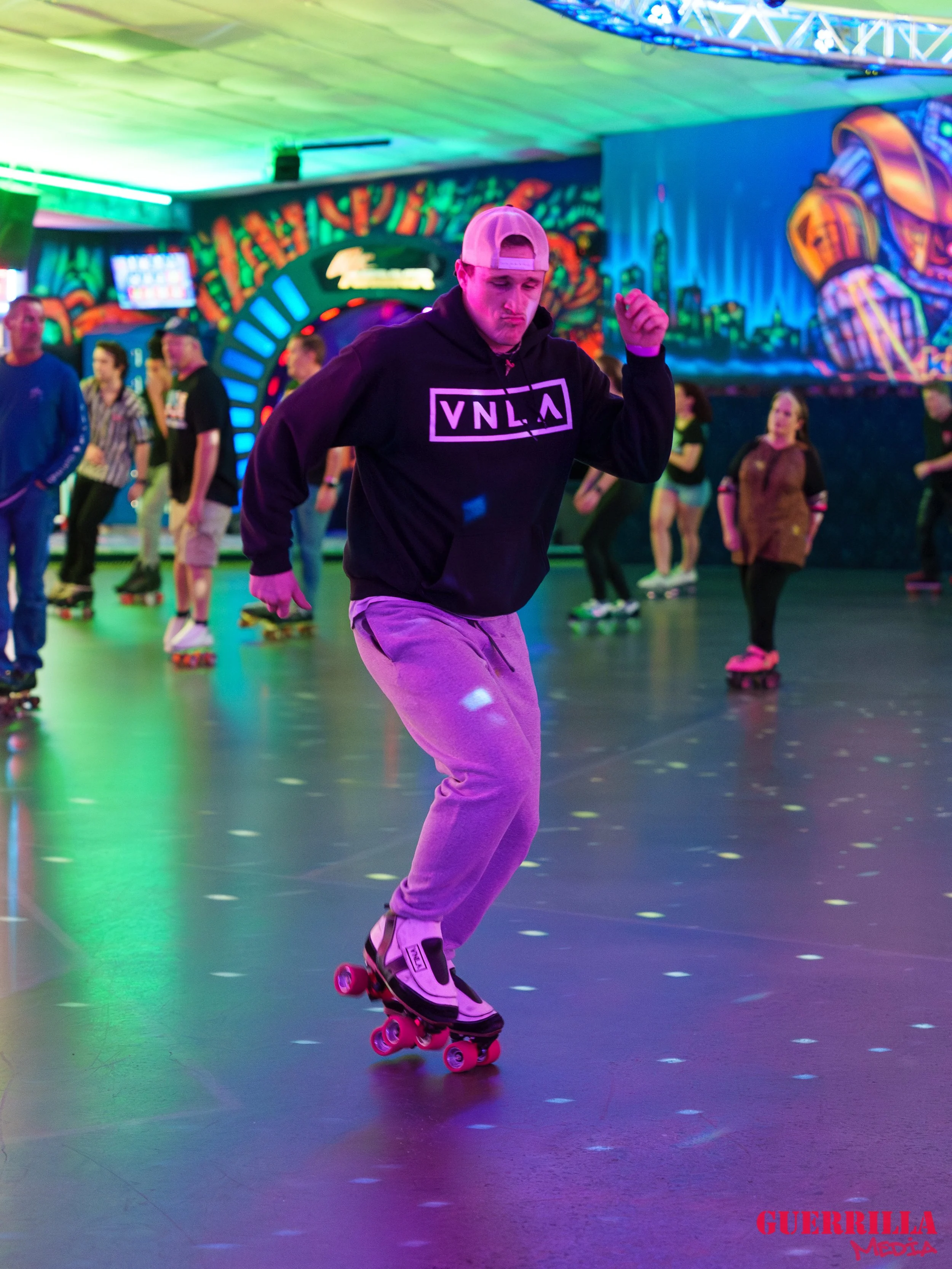 Young man skating at an indoor roller skating rink with colorful lights and murals in the background.