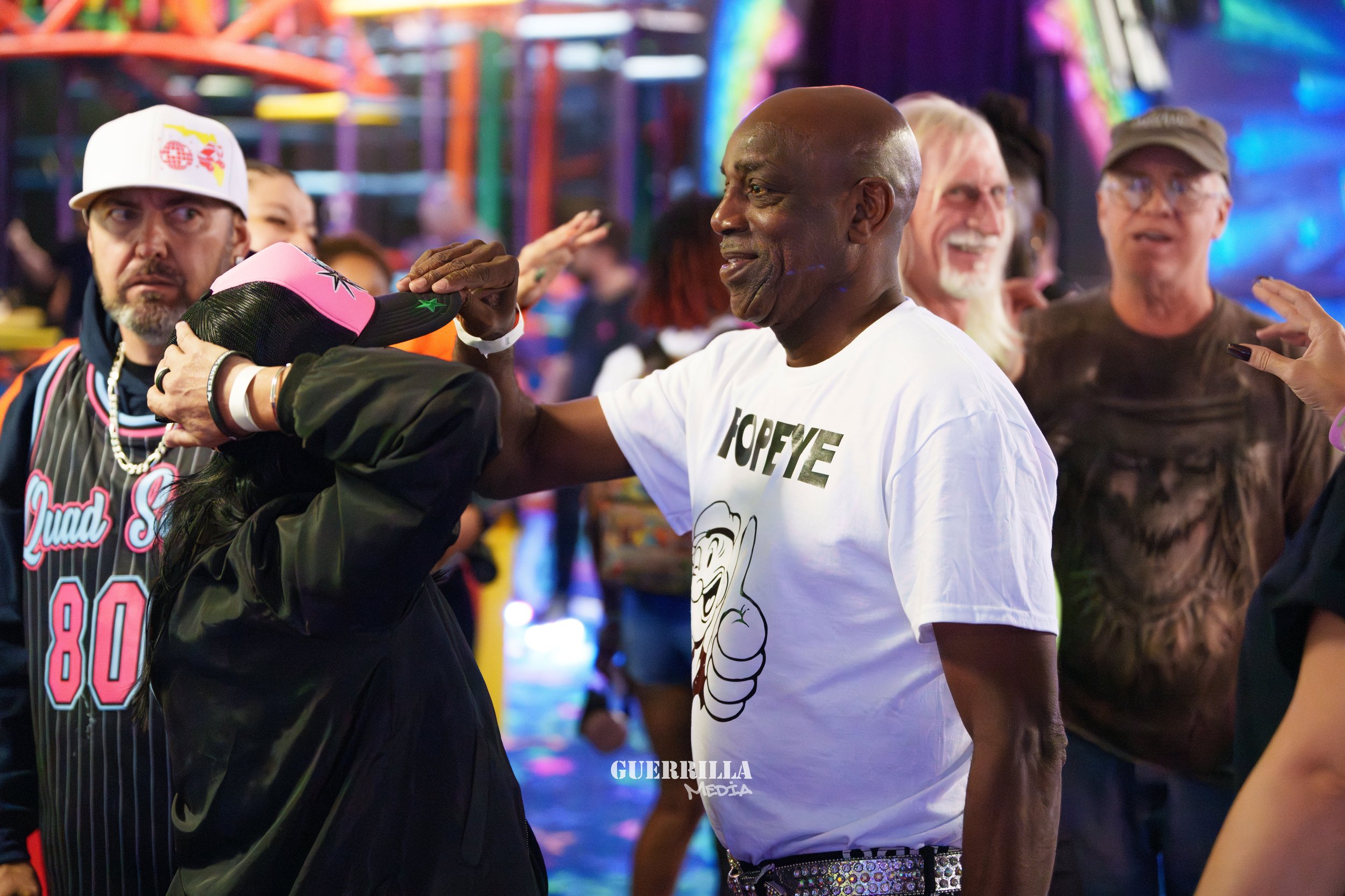 A group of people at a lively event, with a woman wearing a pink and black cap and a black jacket, and a man in a white T-shirt with a graphic and the word 'HOPEYE,' smiling as the woman adjusts his cap. In the background, several other people are en