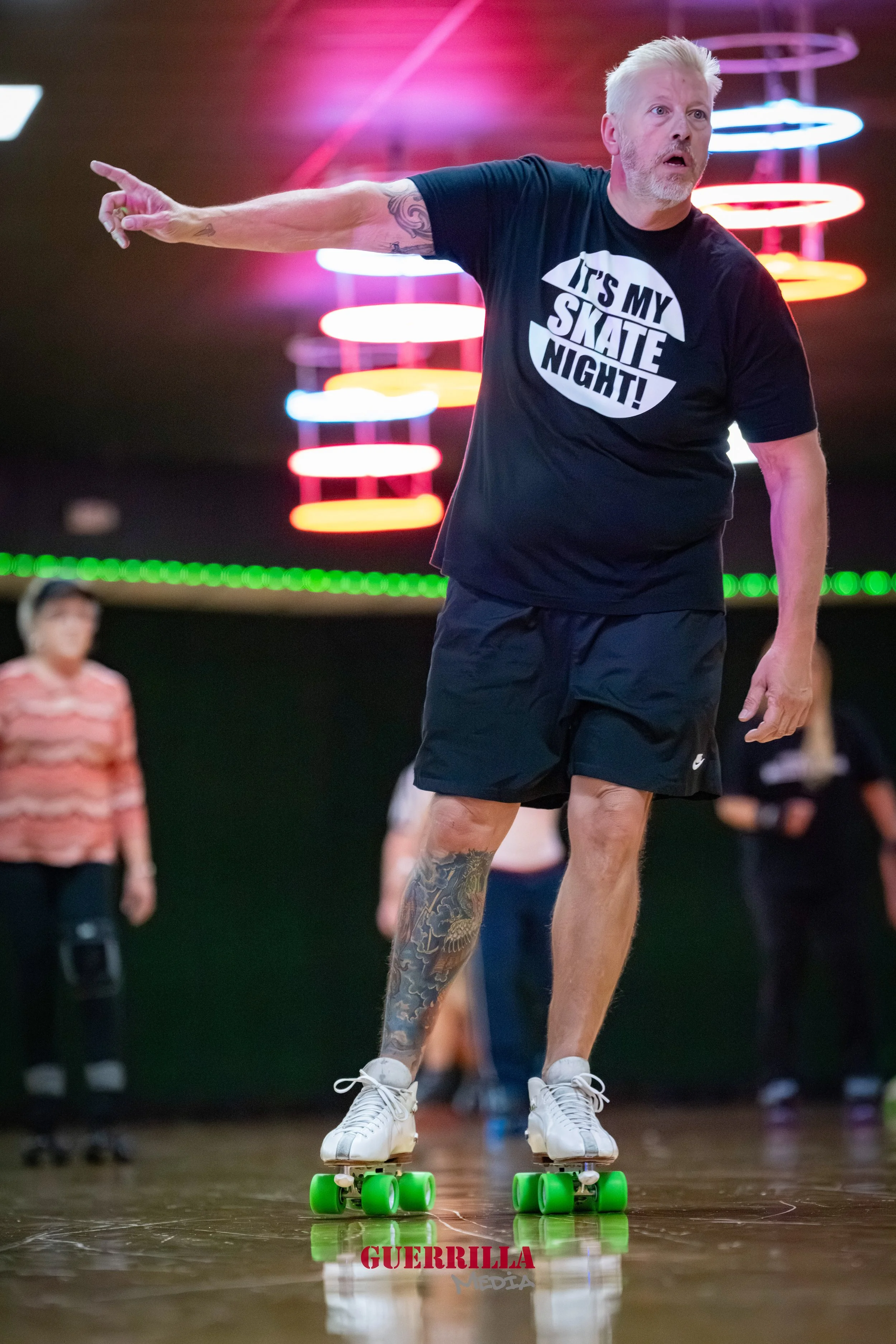 An older man with blonde hair, beard, and tattoos on his left arm and right leg, riding a green-wheeled skateboard indoors. He is wearing a black T-shirt that says 'It’s my skate night!' and black shorts. He appears to be instructing or directing as 