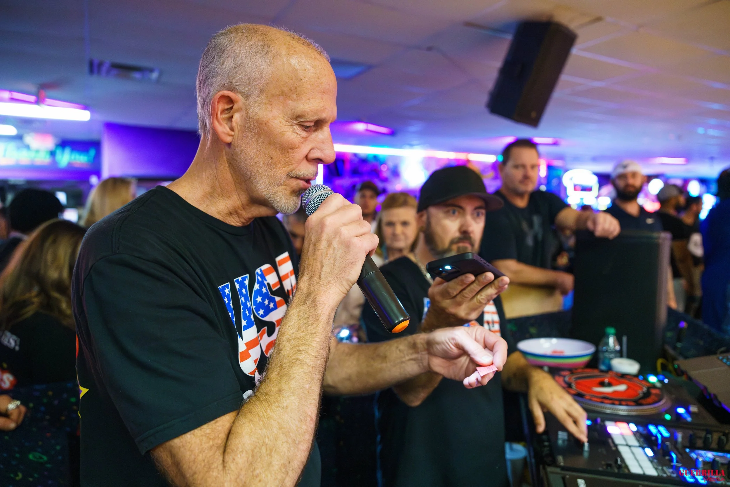 An elderly man with short gray hair and a beard, wearing a black t-shirt with patriotic USA colors, is holding a microphone and reading from a smartphone at a crowded indoor event with colorful neon lights in the background.