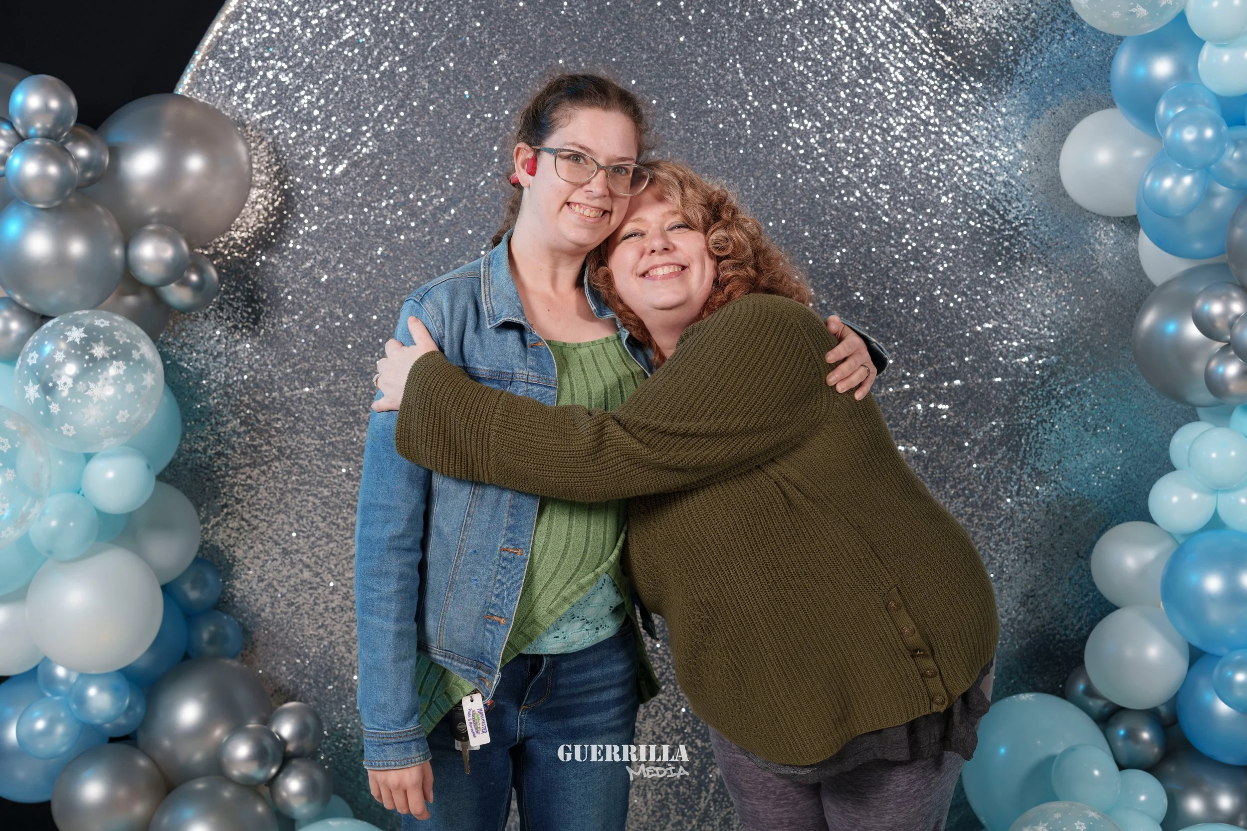 Two women hugging in front of a silver and blue balloon backdrop, smiling at the camera.