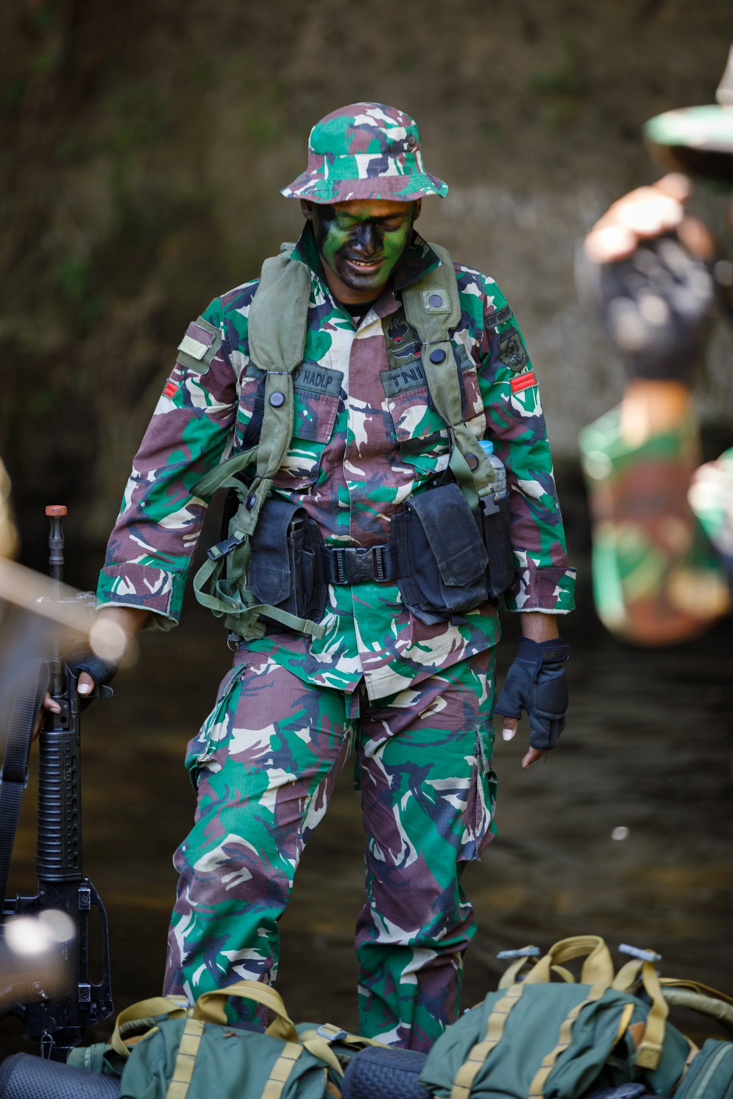 Soldier in camouflage uniform with face paint, holding a rifle, standing near water with backpacks in the foreground.