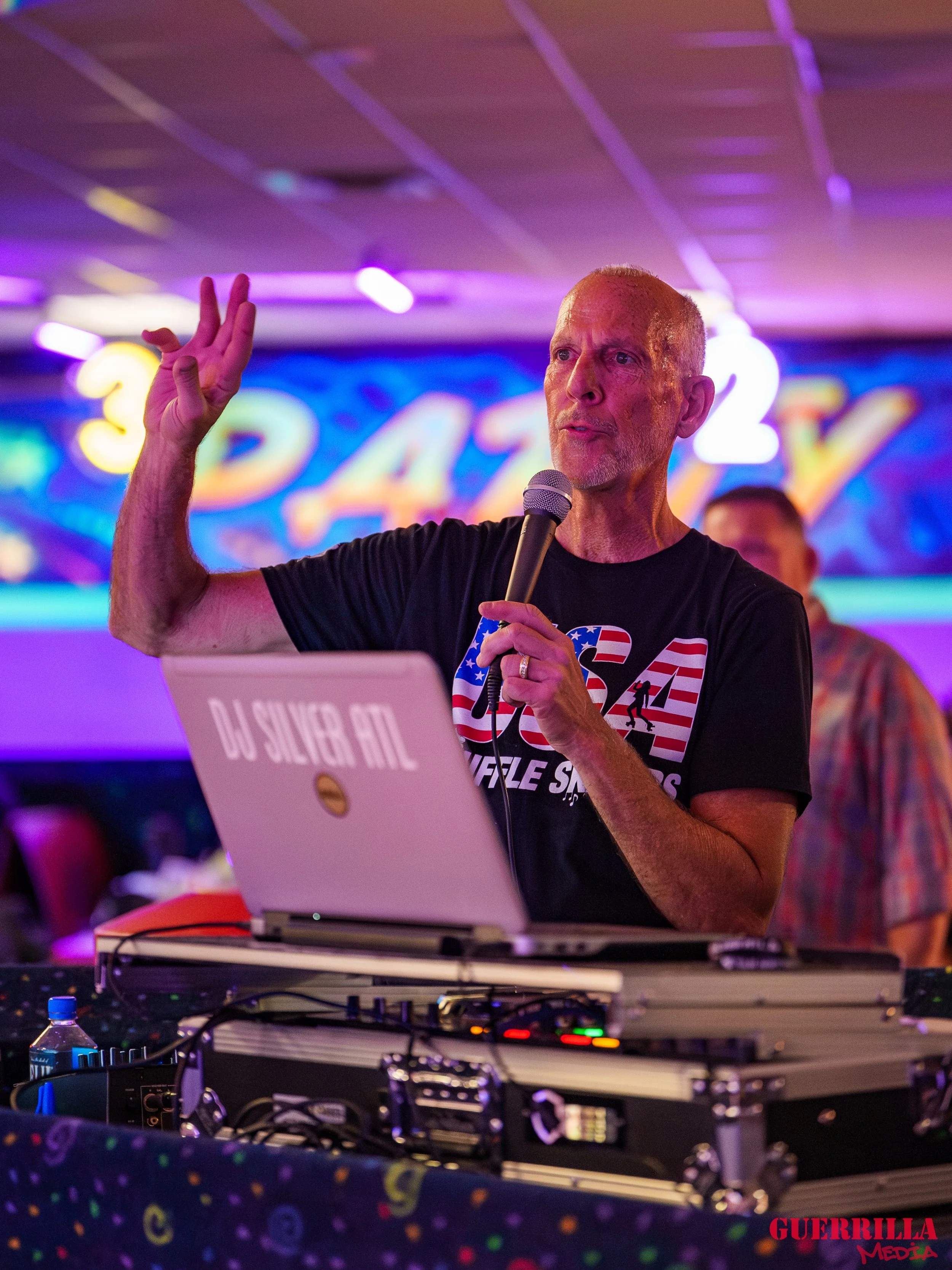 A man with short gray hair and a goatee speaking into a microphone, standing behind DJ equipment with a laptop labeled 'DJ SILVER PUB.' He is wearing a black T-shirt with an American flag design and a logo. The background features colorful neon light