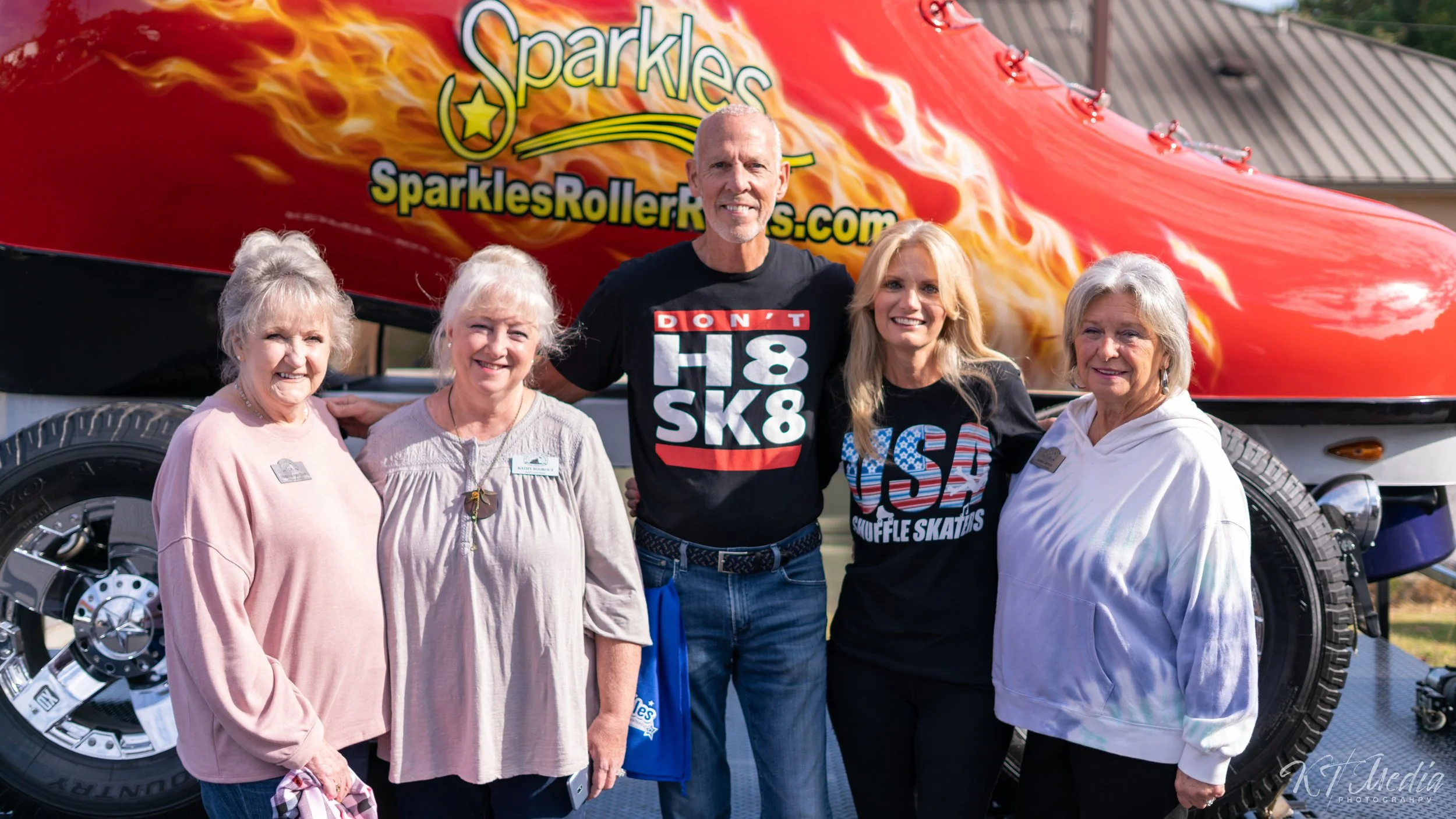 Group of five people standing in front of a red and black roller coaster with the sign 'Sparkles' and 'SparklesRollerRinks.com' in the background. The group includes four women and one man, all smiling at the camera.