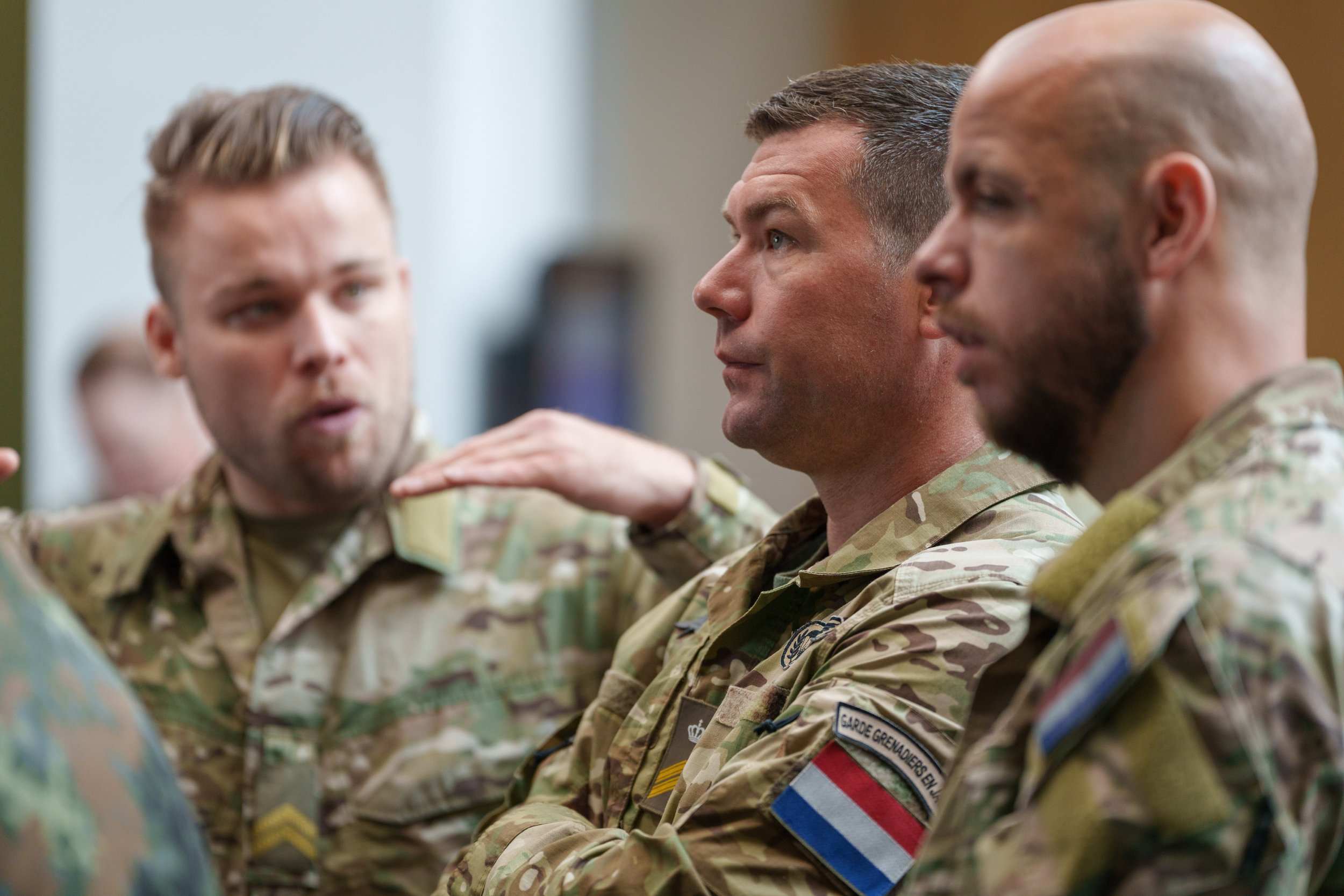Three military personnel in camouflage uniforms engaged in a discussion indoors, with one person explaining and the other listening.