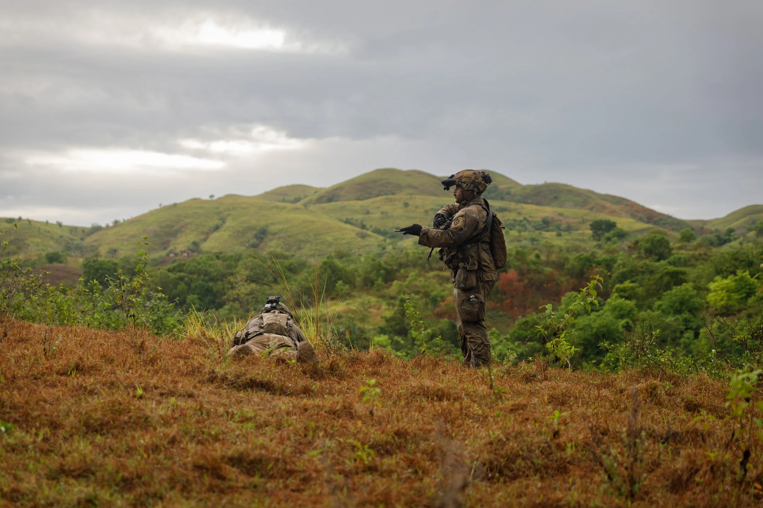 Two soldiers in tactical gear conduct training or patrol in a hilly, grassy landscape during overcast weather.