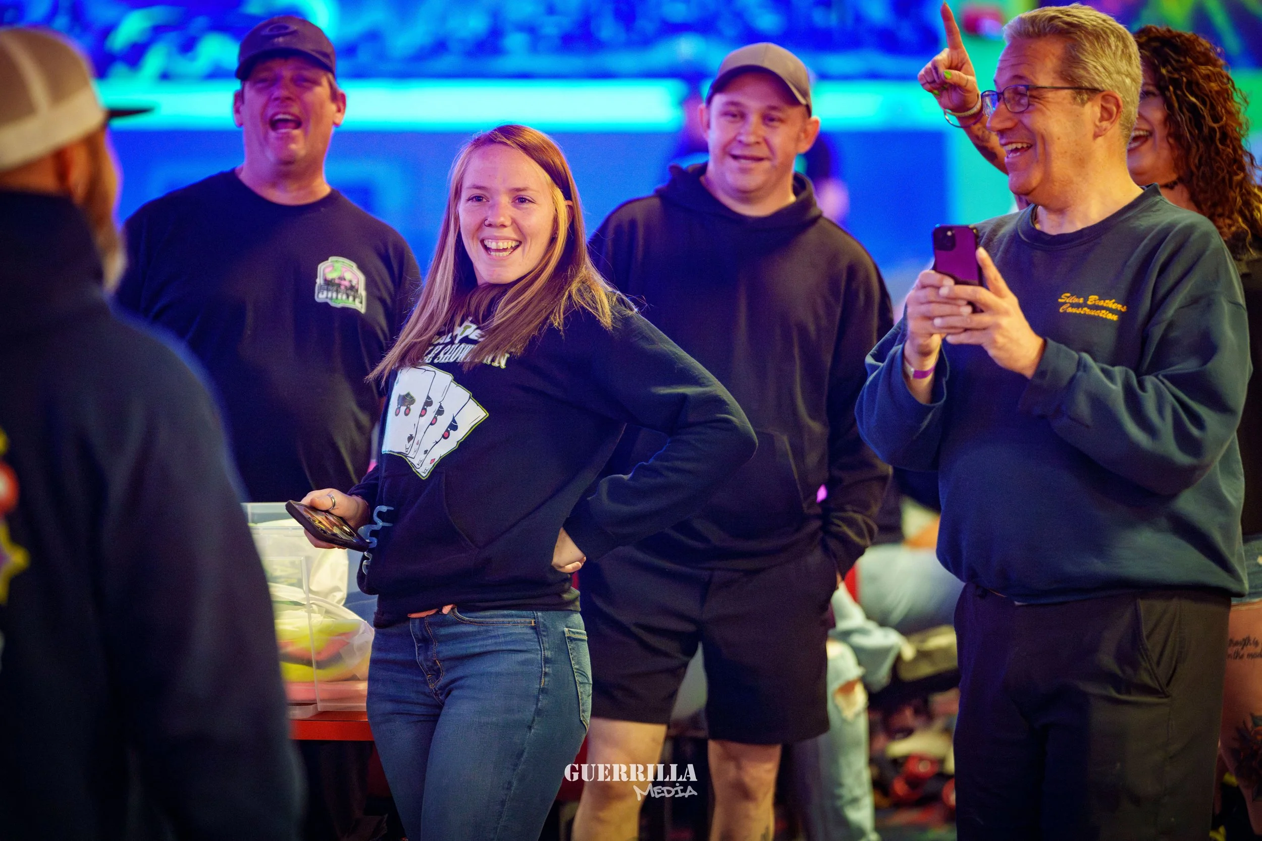 A group of people laughing and smiling at an indoor event with colorful lighting, some holding smartphones, including a young woman in a black hoodie and a man with glasses in a green sweatshirt.