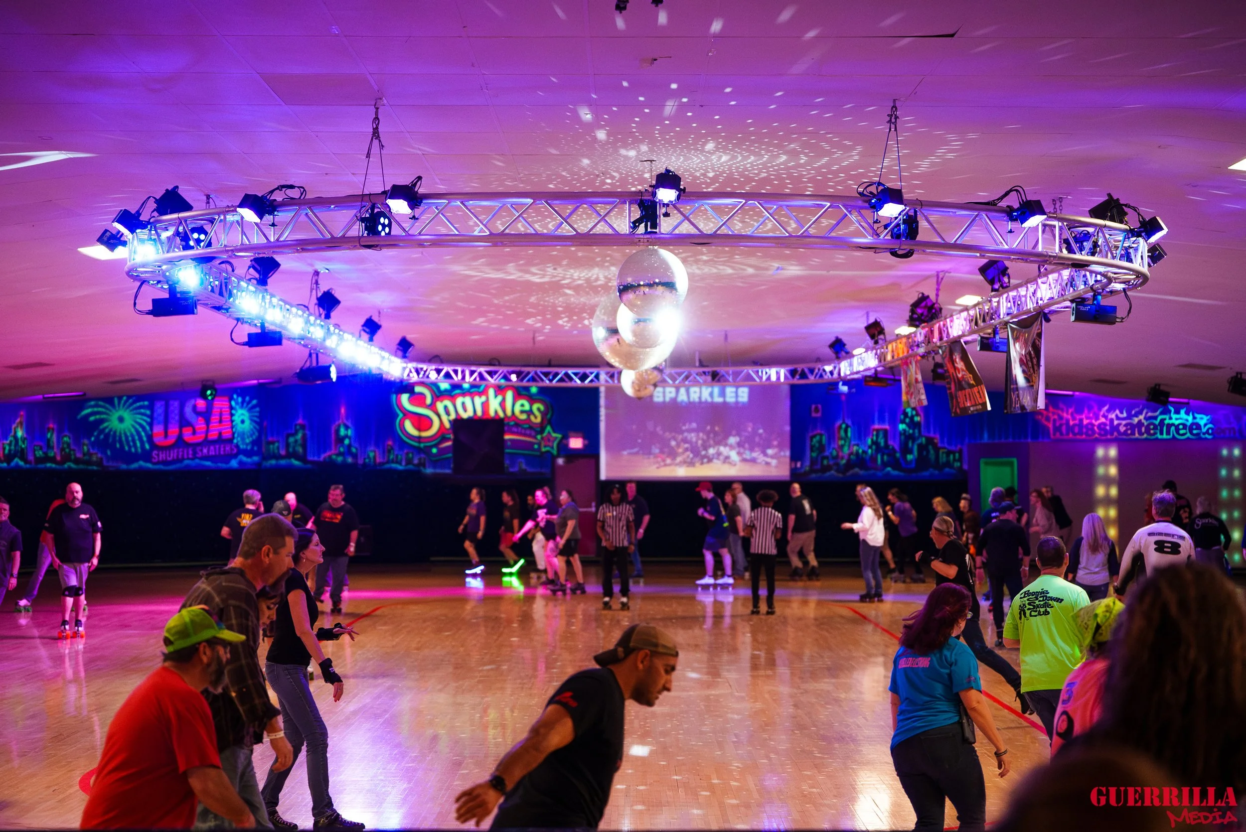 People roller skating on a brightly lit indoor roller rink with colorful neon signs and a disco ball overhead.