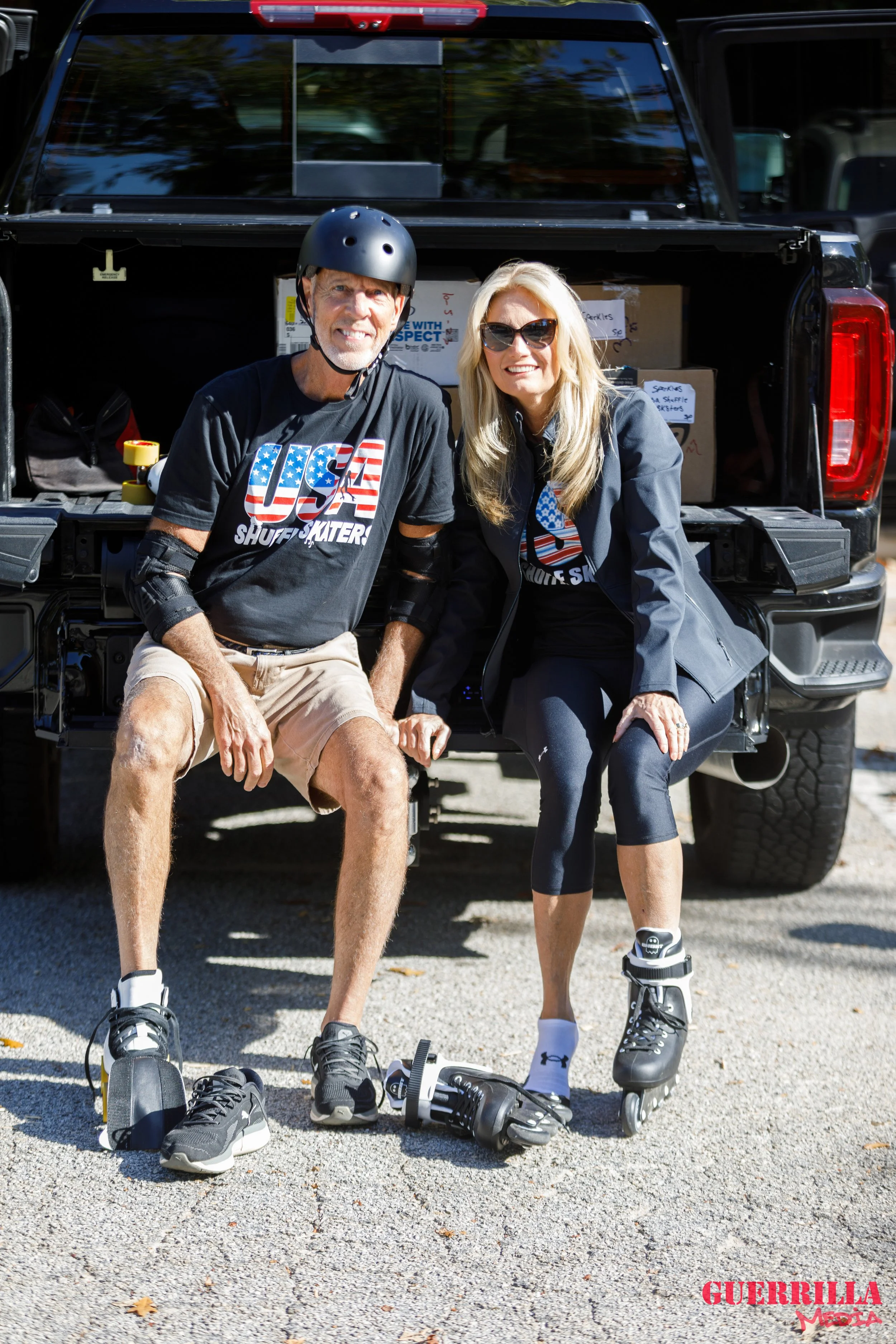 Two people sitting on the back of a pickup truck, wearing roller skates and protective gear, smiling at the camera.