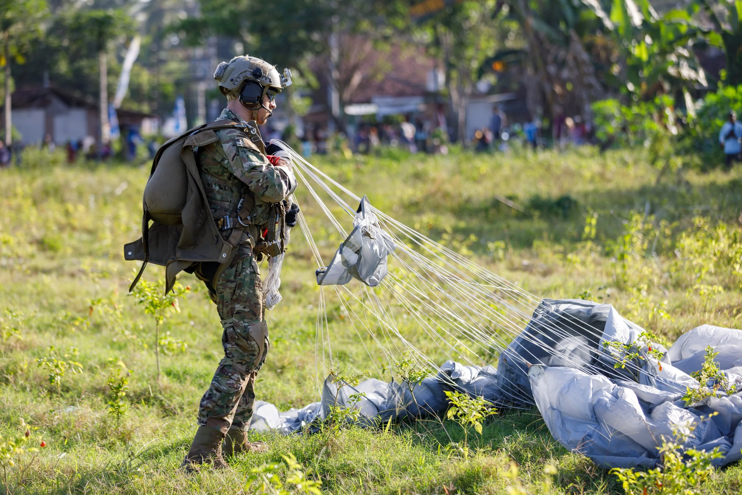A soldier in camouflage uniform, wearing a helmet and backpack, is standing on grass in a field, handling a large shadow parachute with lines attached. In the background, there are trees, buildings, and people.