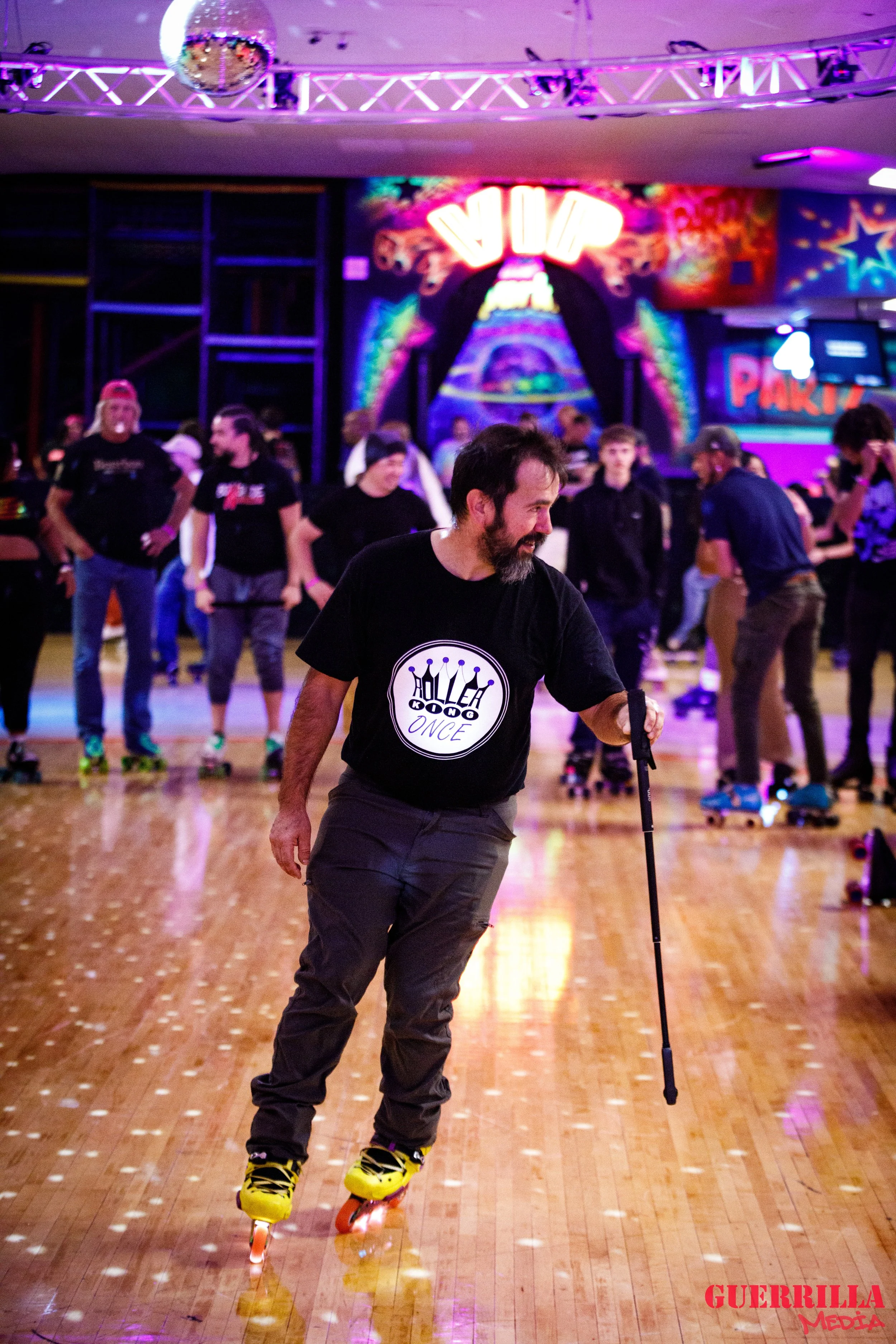A man with a beard rollerblading on a wooden roller rink, holding a cane, at a lively indoor roller skating rink with neon lights and colorful decorations.