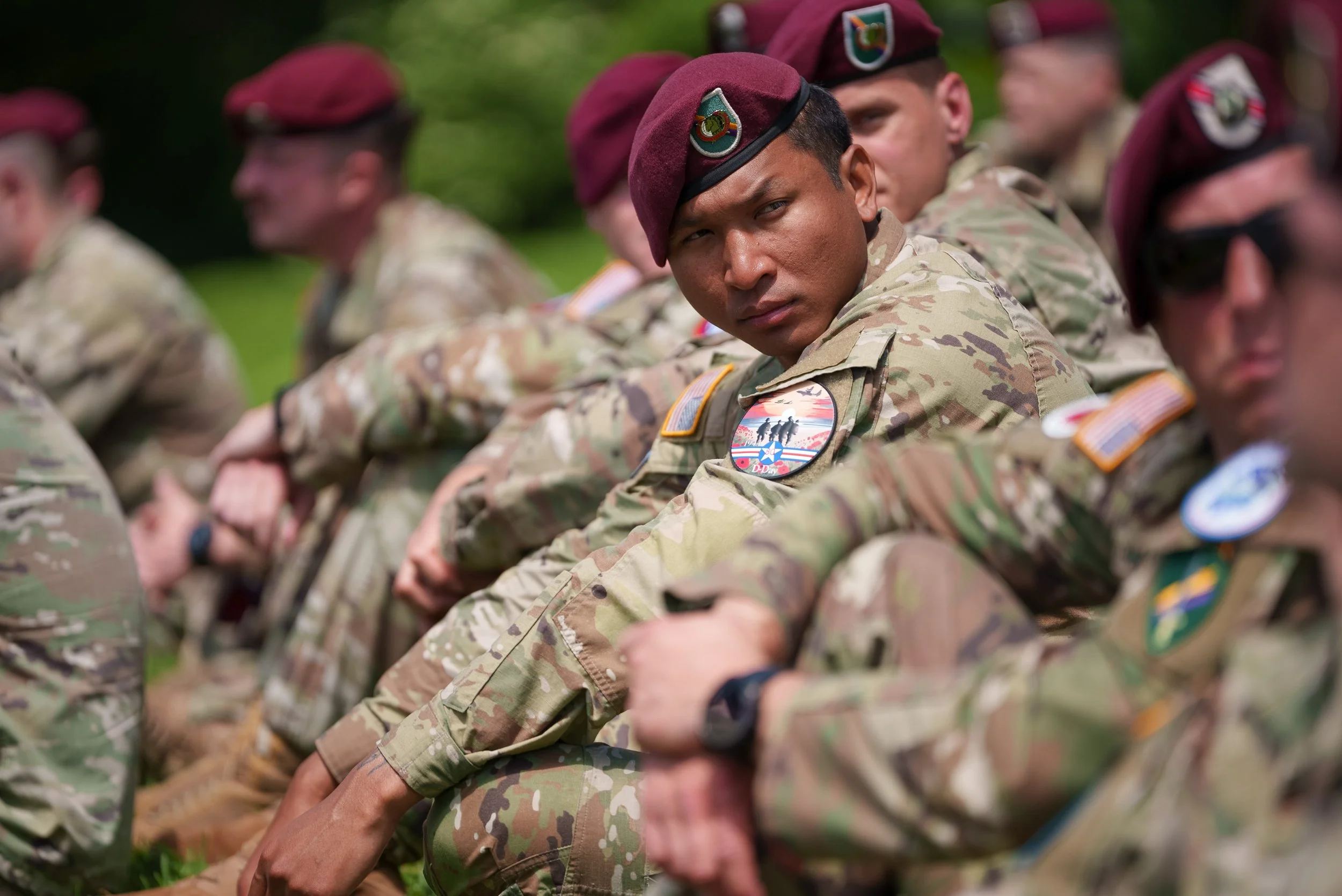 Group of soldiers sitting outdoors, wearing camouflage uniforms and maroon berets, with one soldier looking directly at the camera.