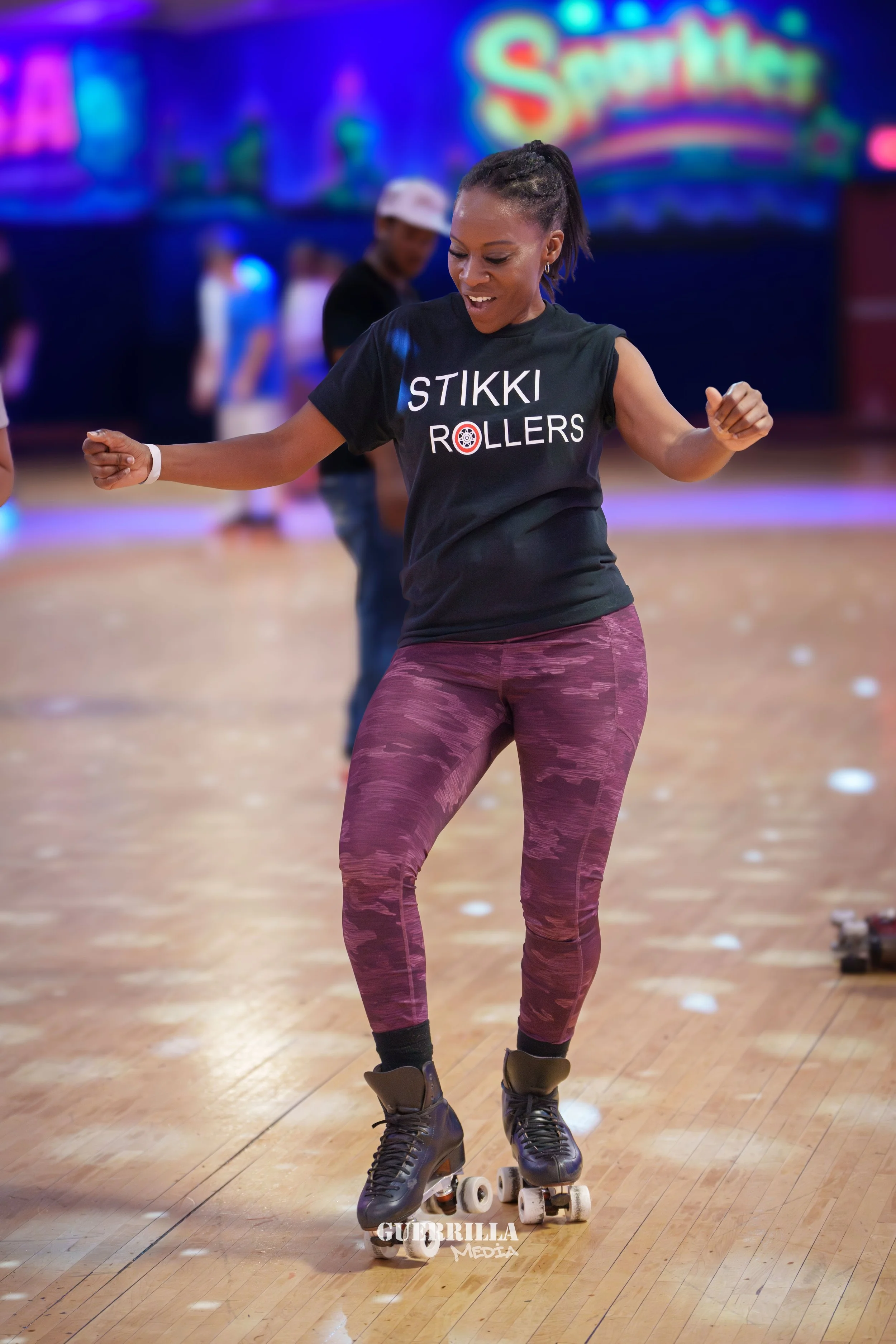 A woman roller-skating indoors, wearing a black T-shirt with 'STIKKI ROLLERS' printed on it, purple camouflage leggings, and black roller skates, with a smile on her face.