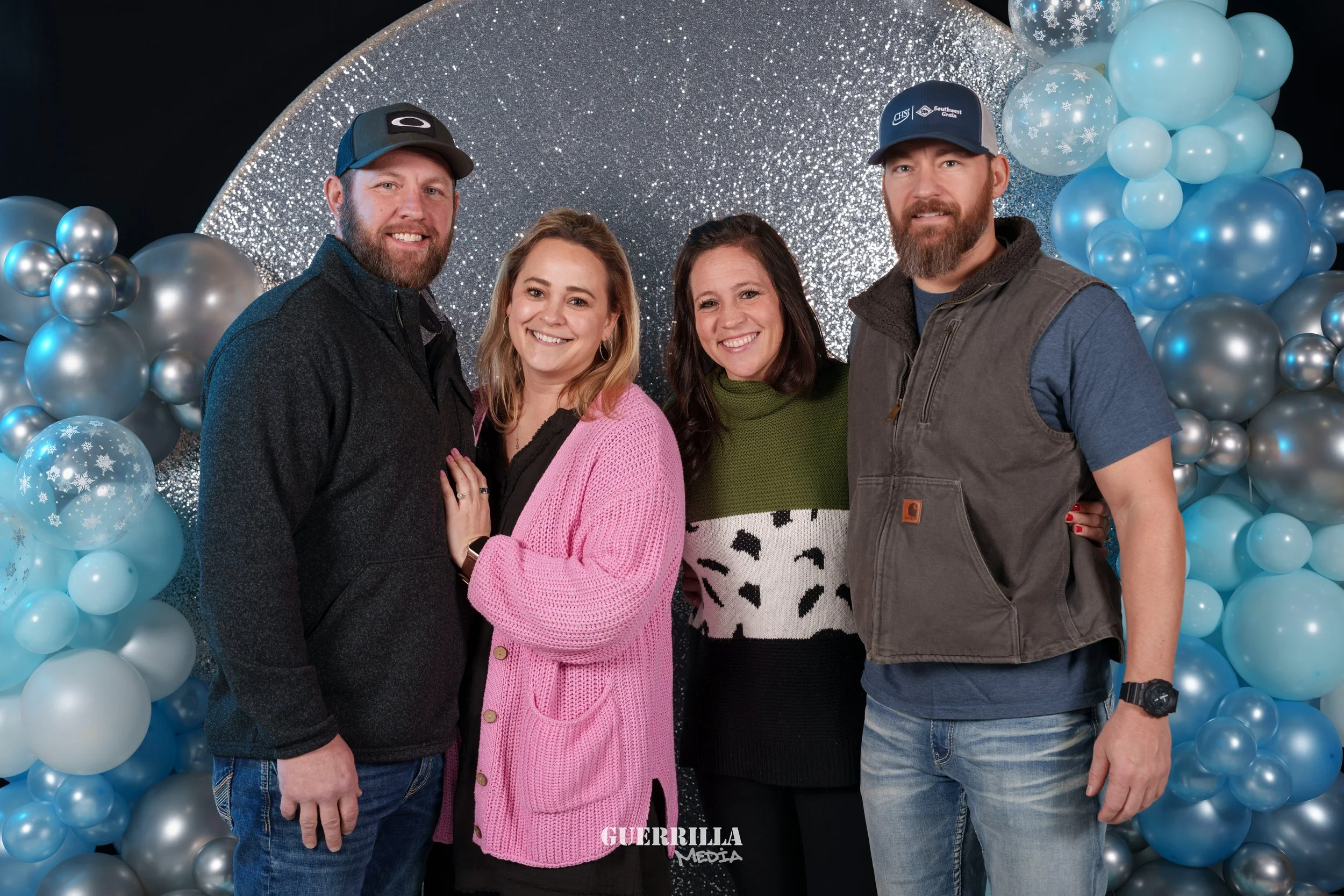 Four people smiling and standing close together in front of a silver and blue balloon backdrop. Two men and two women, dressed casually.