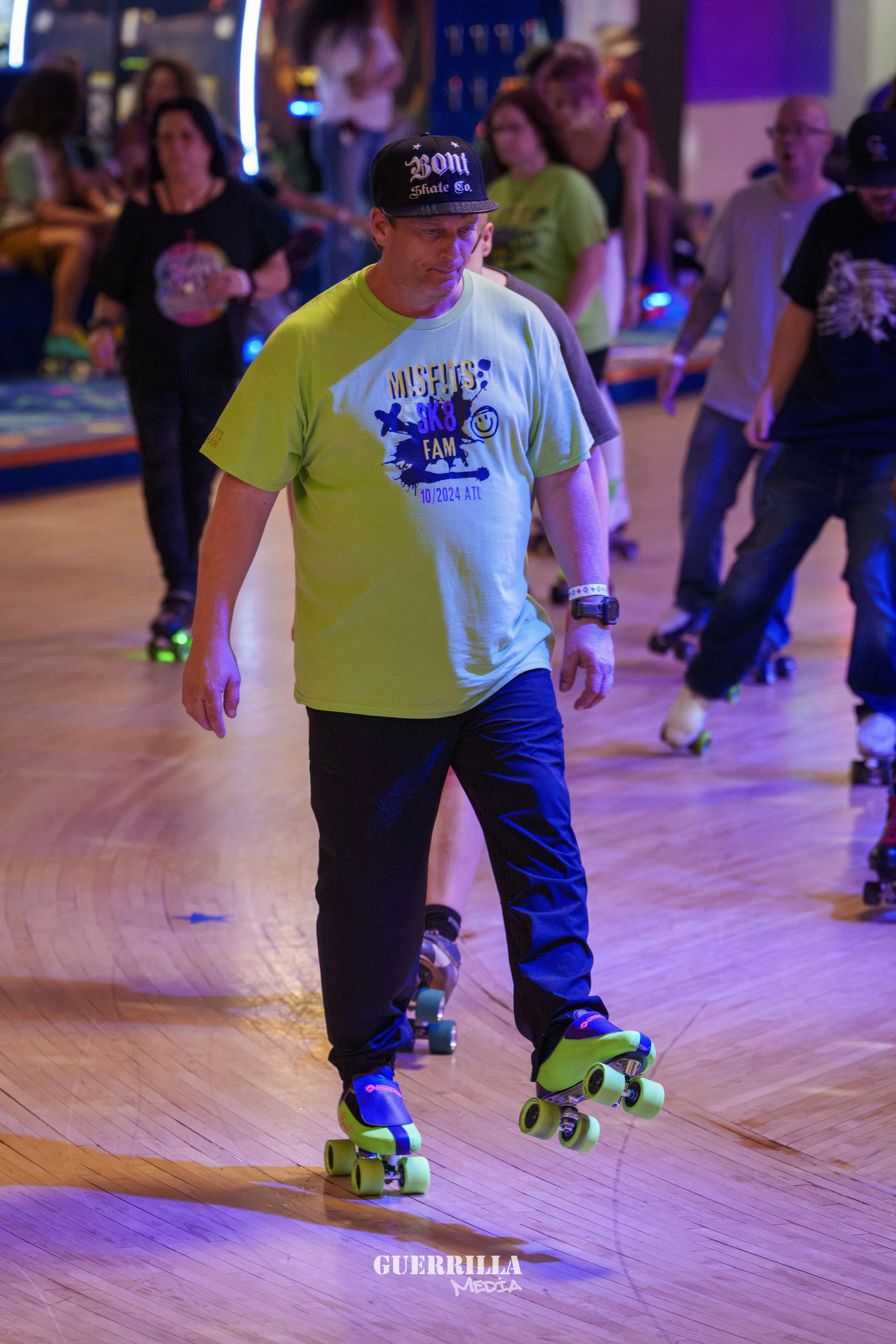 Man in lime green T-shirt and black pants roller skating indoors, with other skaters and spectators in the background.