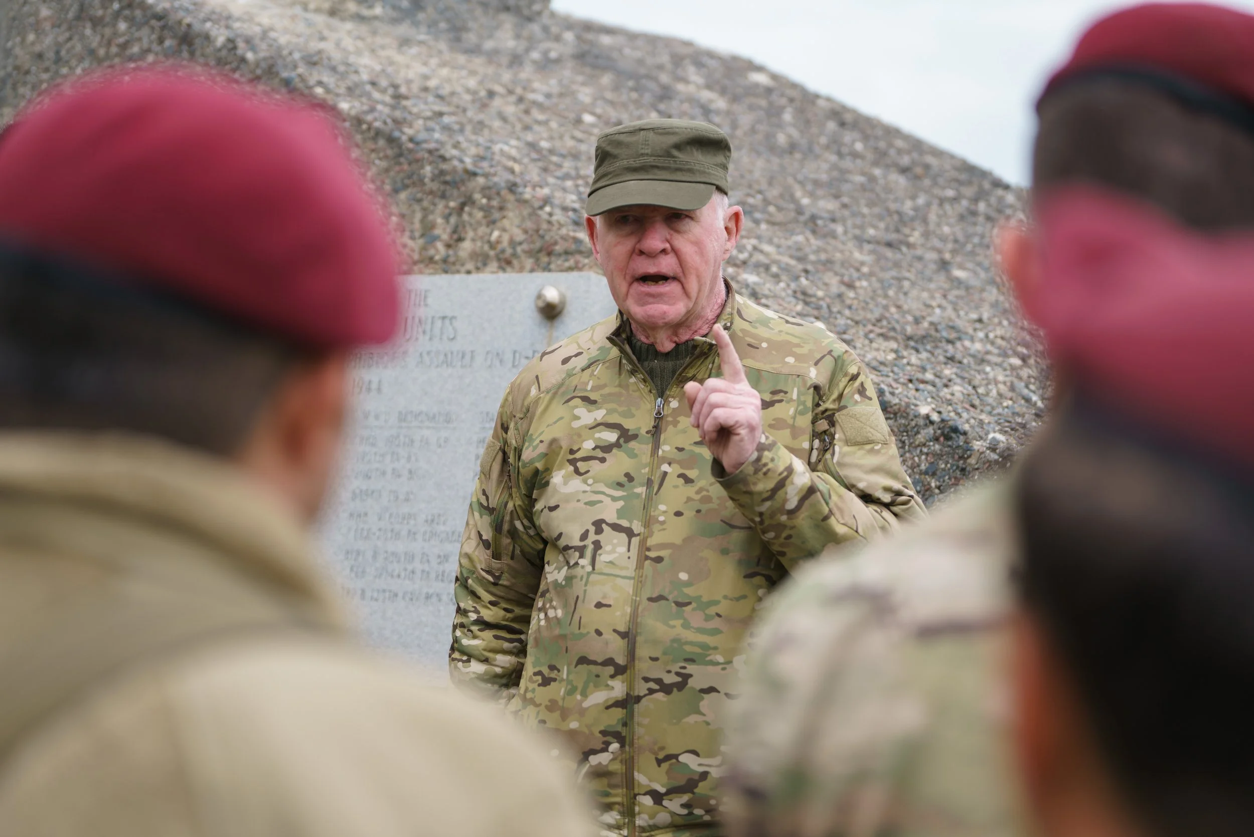 A military officer in camouflage uniform speaking to a group of soldiers outdoors near a memorial.