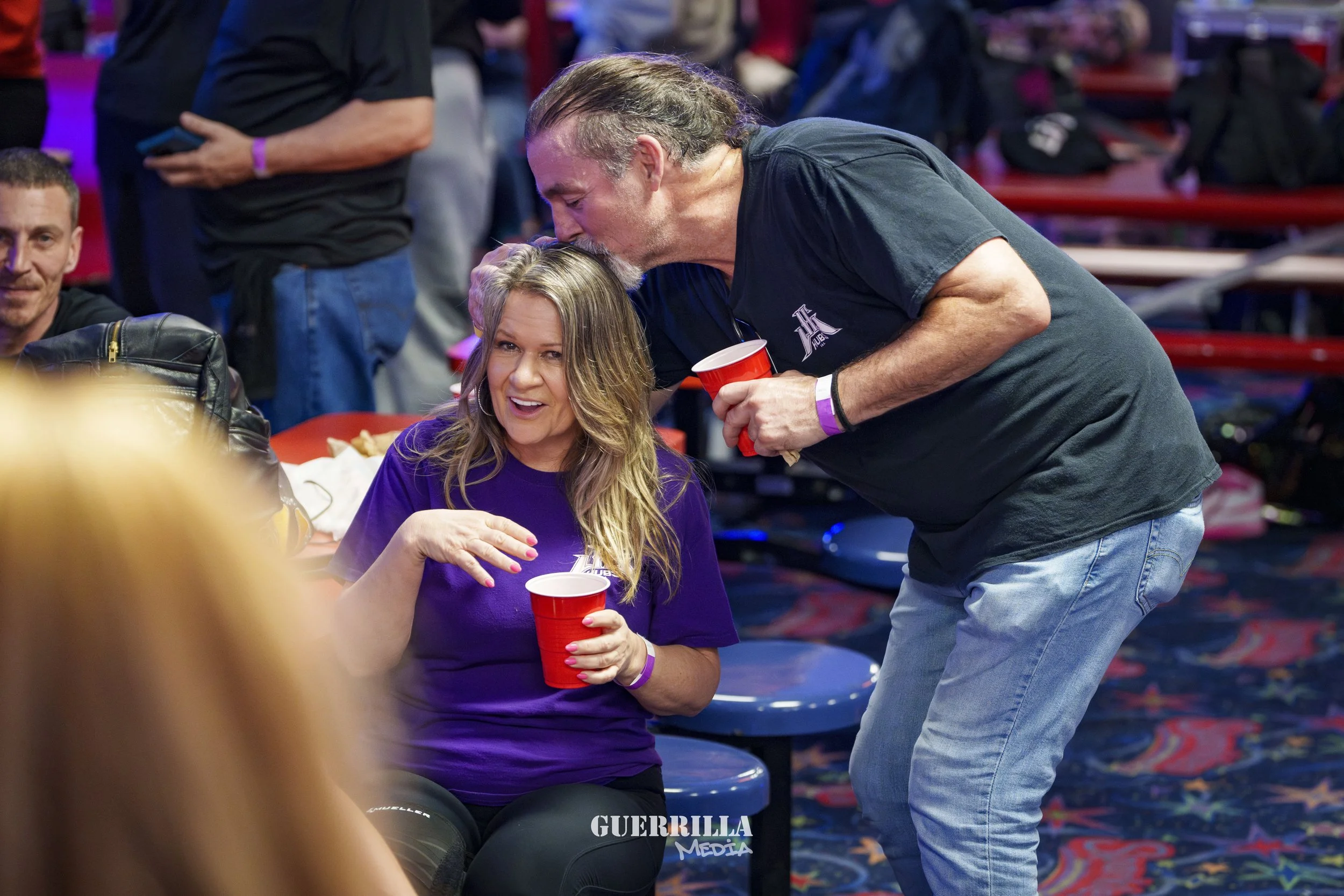 A woman in a purple shirt sitting on a blue seat holding a red cup, as a man with long gray hair kissing her forehead at a social event with other people in the background.