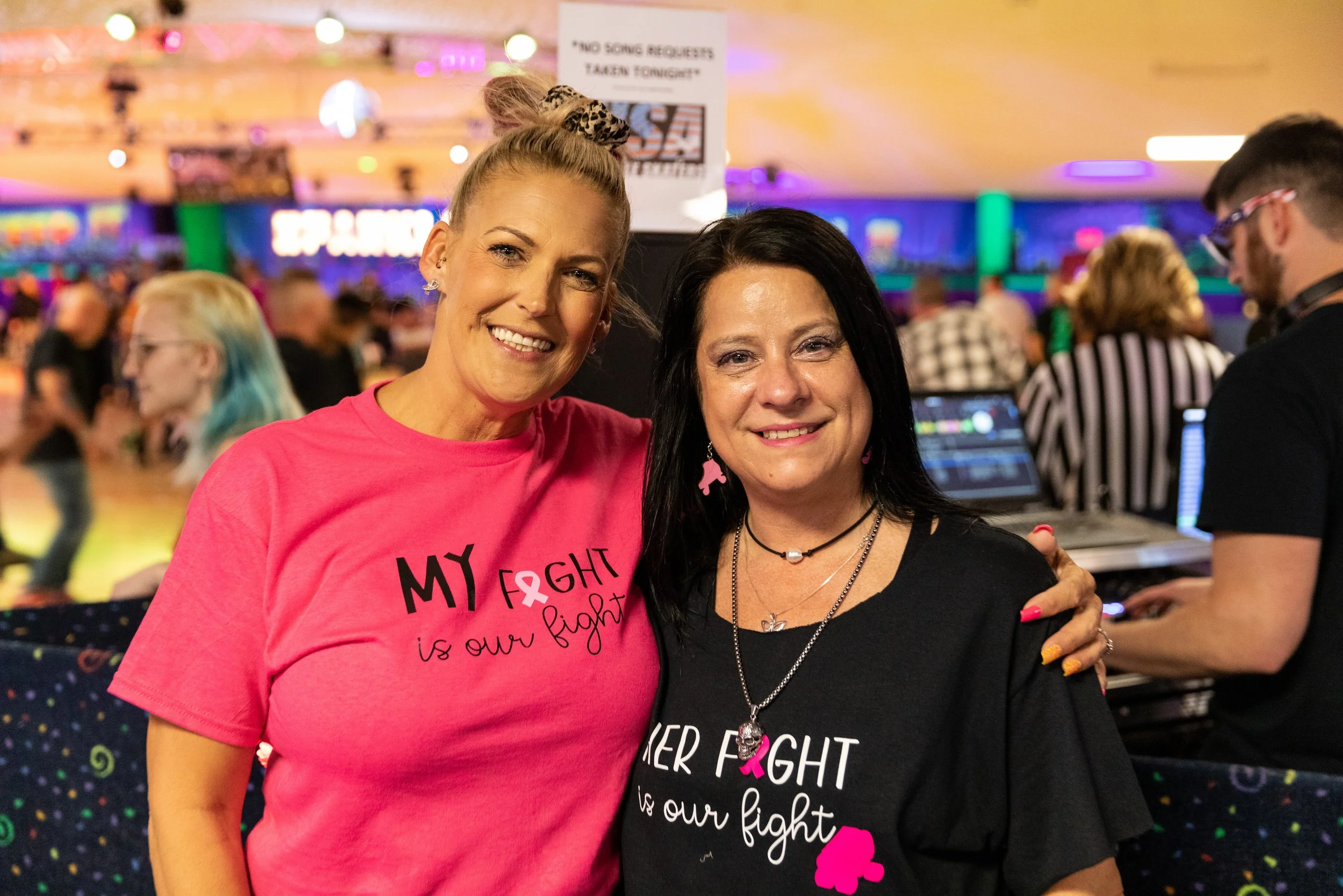 Two women smiling and posing together at an indoor event with colorful background. They are wearing pink and black T-shirts supporting breast cancer awareness, with messages about fighting cancer.