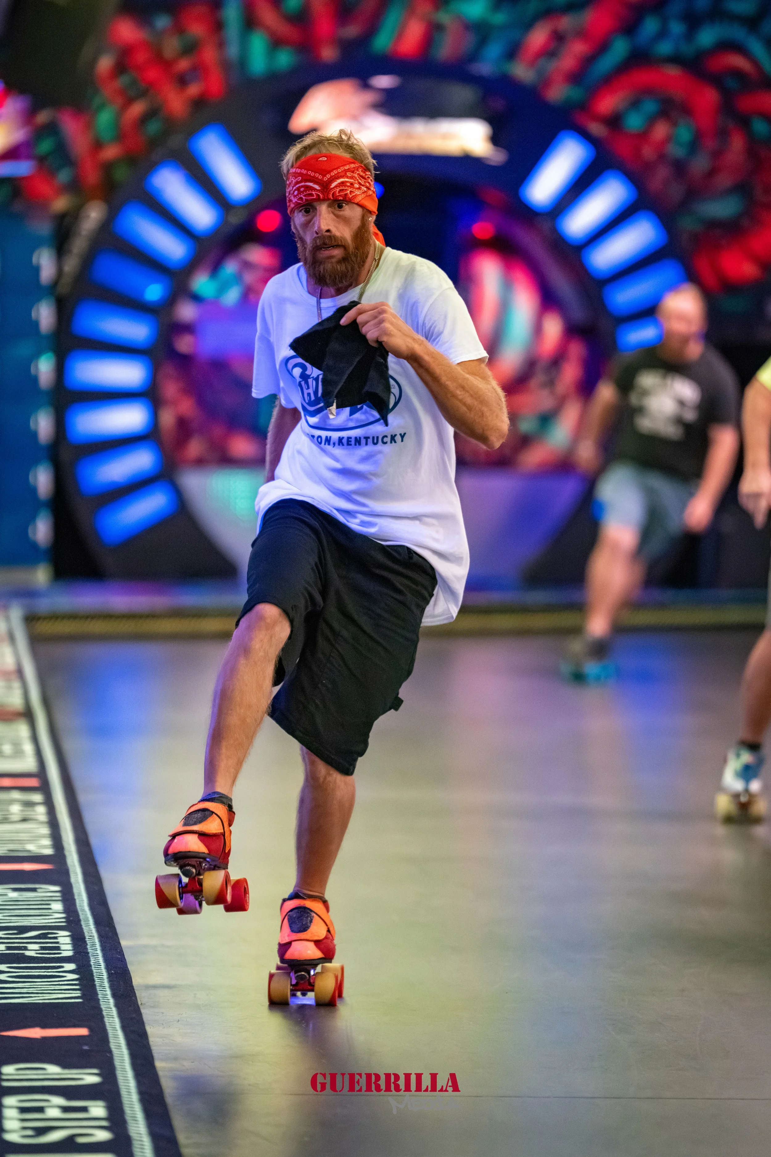Man with a red bandana roller skating indoors, wearing a white T-shirt and black shorts, with a colorful abstract background.