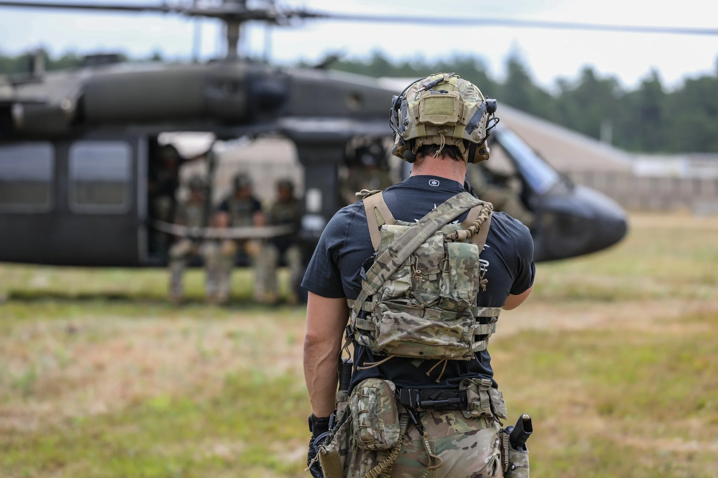 A soldier with camouflage gear and a helmet standing in front of a black helicopter with other soldiers inside, on an open field.