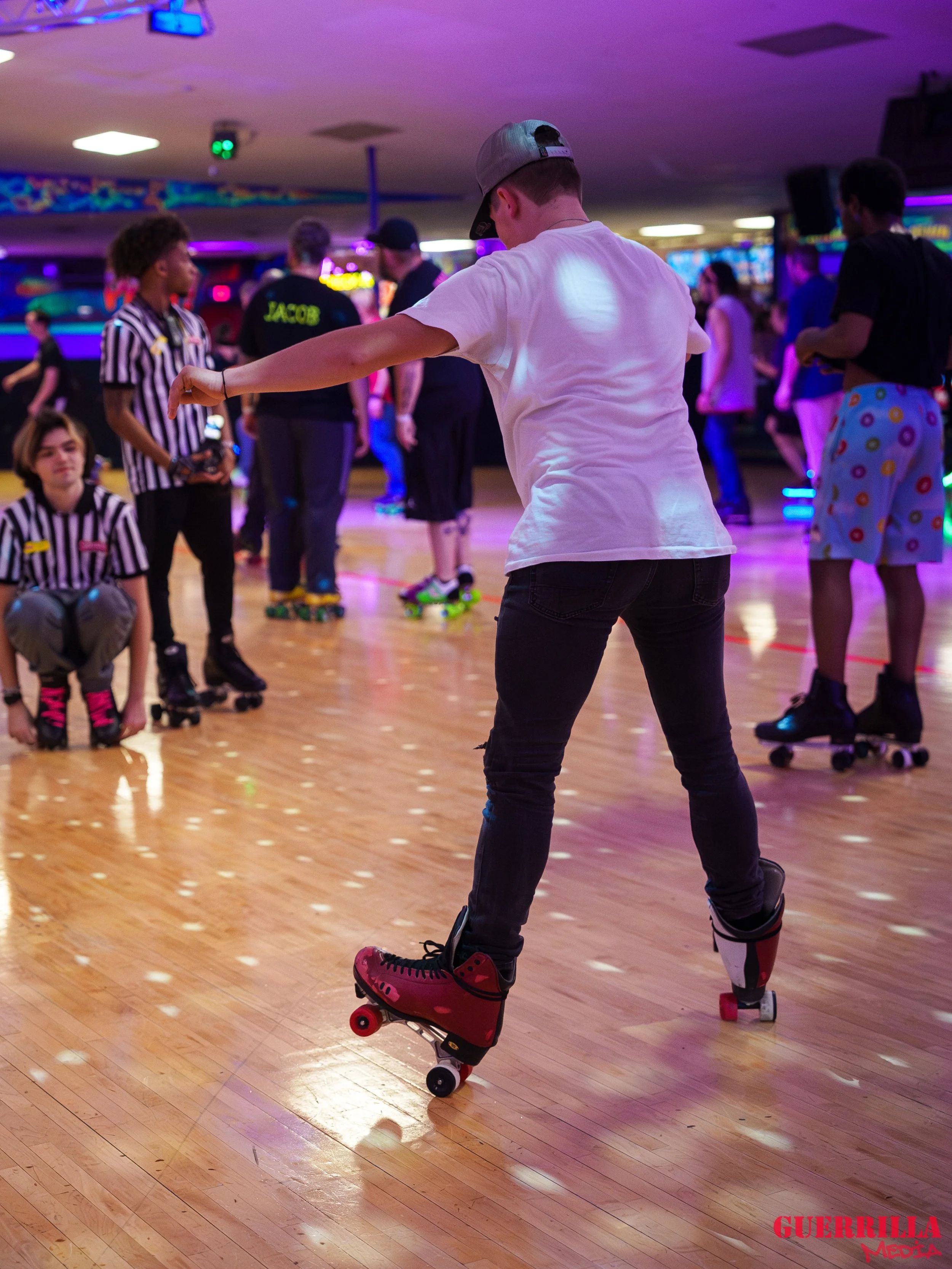 A young person roller skates on a wooden floor at an indoor roller skating rink. They are wearing a white t-shirt, a gray cap, and red sneakers. Other skaters and a skate referee are visible in the background under colorful lights.