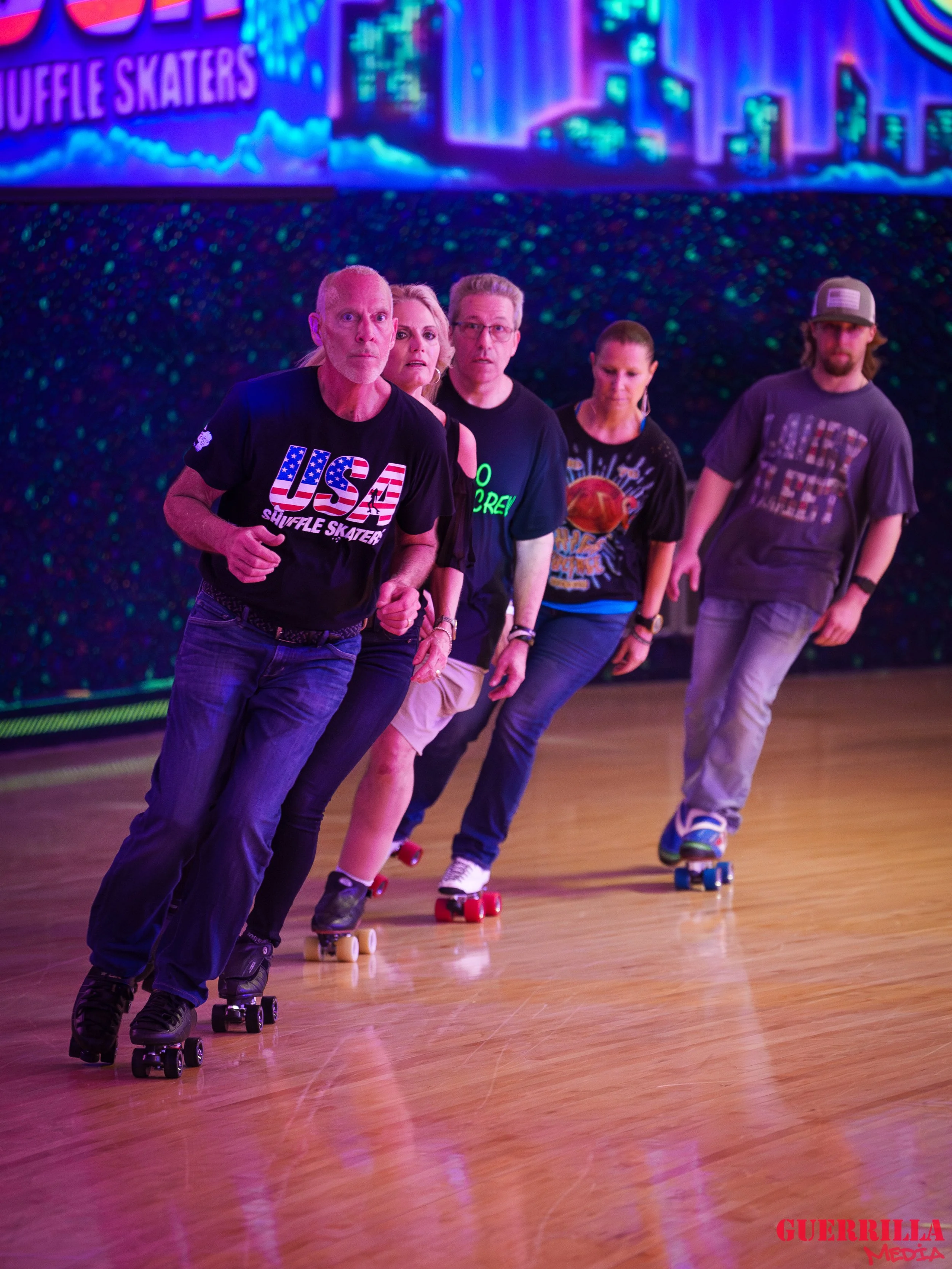 Five people roller skating indoors in a line, with neon colorful background and wooden floor, one person wearing a black USA Shuffle Skaters shirt, others in casual clothes. 
