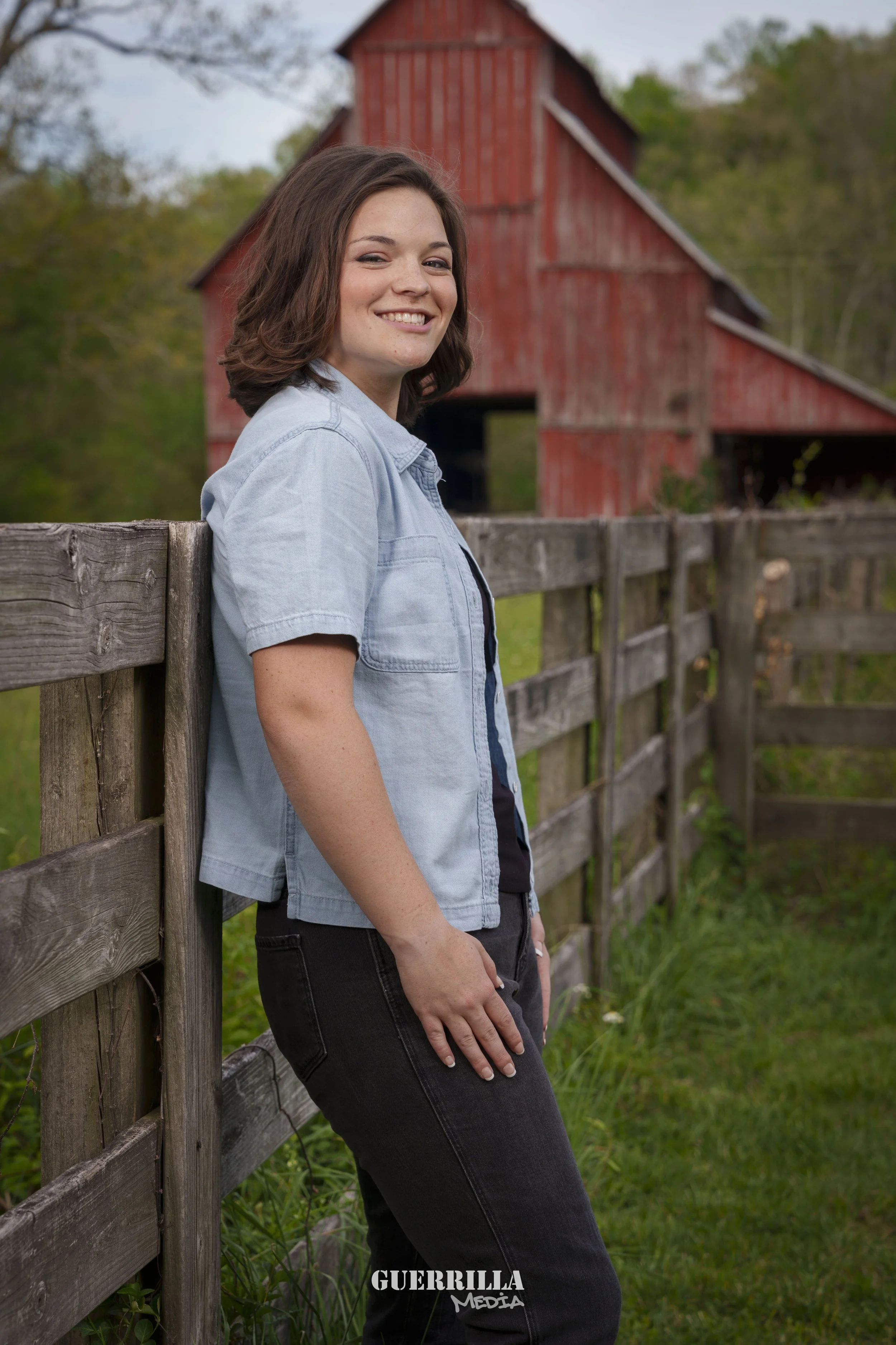 A young woman with shoulder-length brown hair smiling while leaning against a wooden fence. She is wearing a light blue denim jacket over a dark shirt, with black pants. In the background, there is a red barn surrounded by green trees and grass.