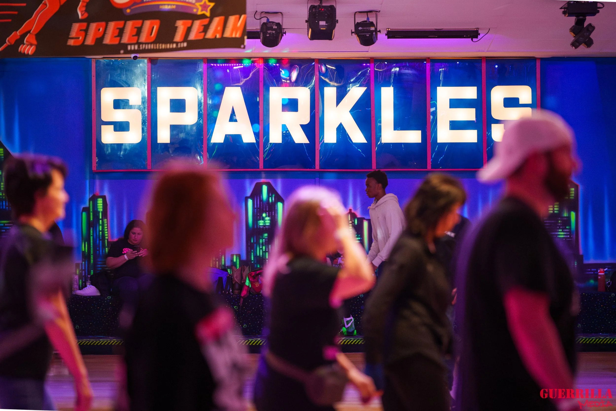 People roller skating in an indoor rink with colorful neon lights and a large illuminated sign that reads 'SPARKLES' in the background.
