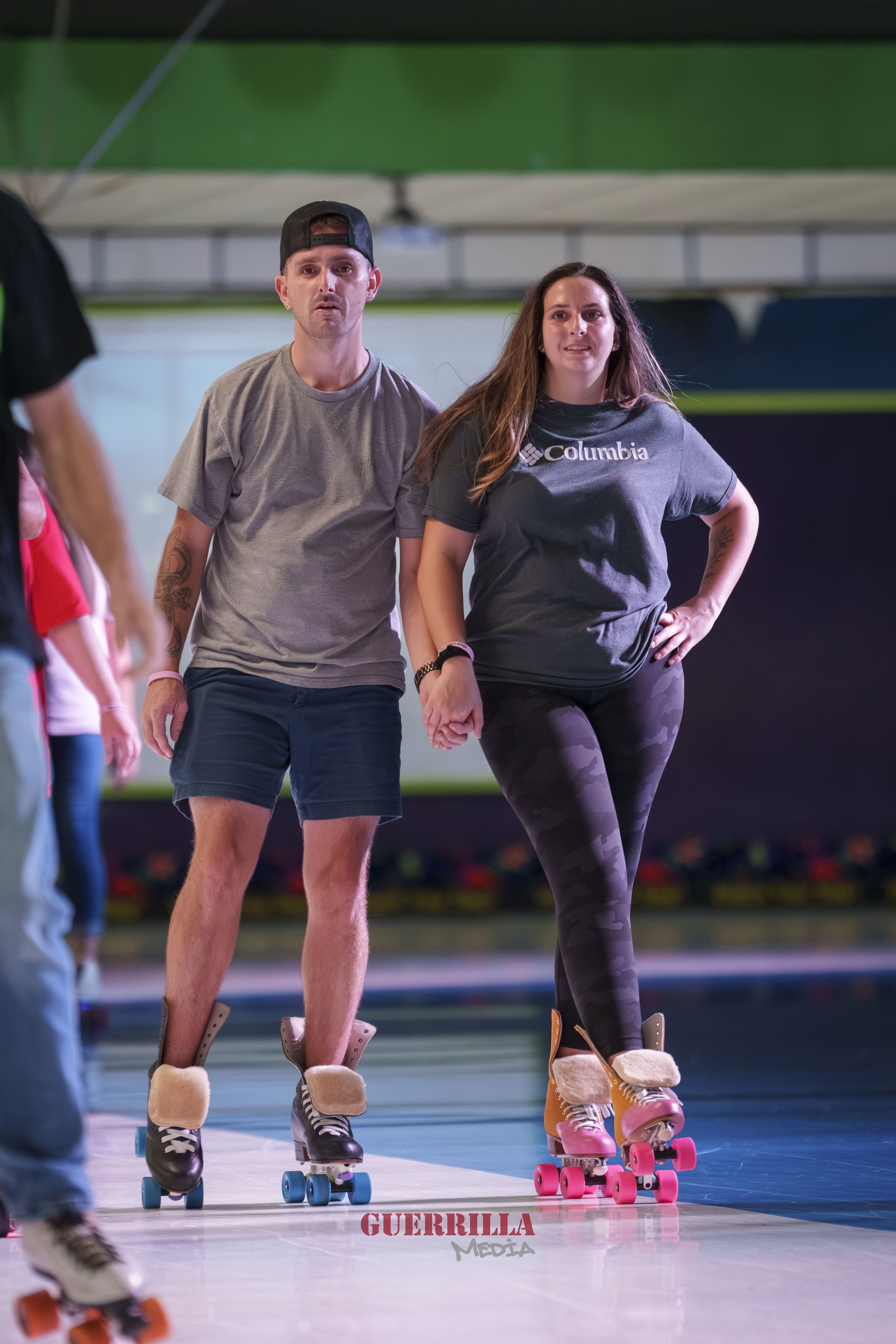 A man and woman roller skating together indoors, holding hands, with the woman wearing a Columbia t-shirt and camouflage leggings.