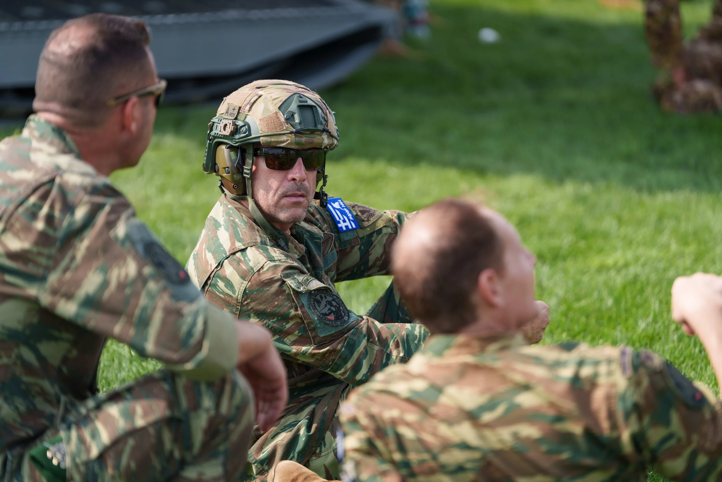 Military personnel sitting on grass during training or break, wearing camouflage uniforms, tactical gear, and sunglasses.