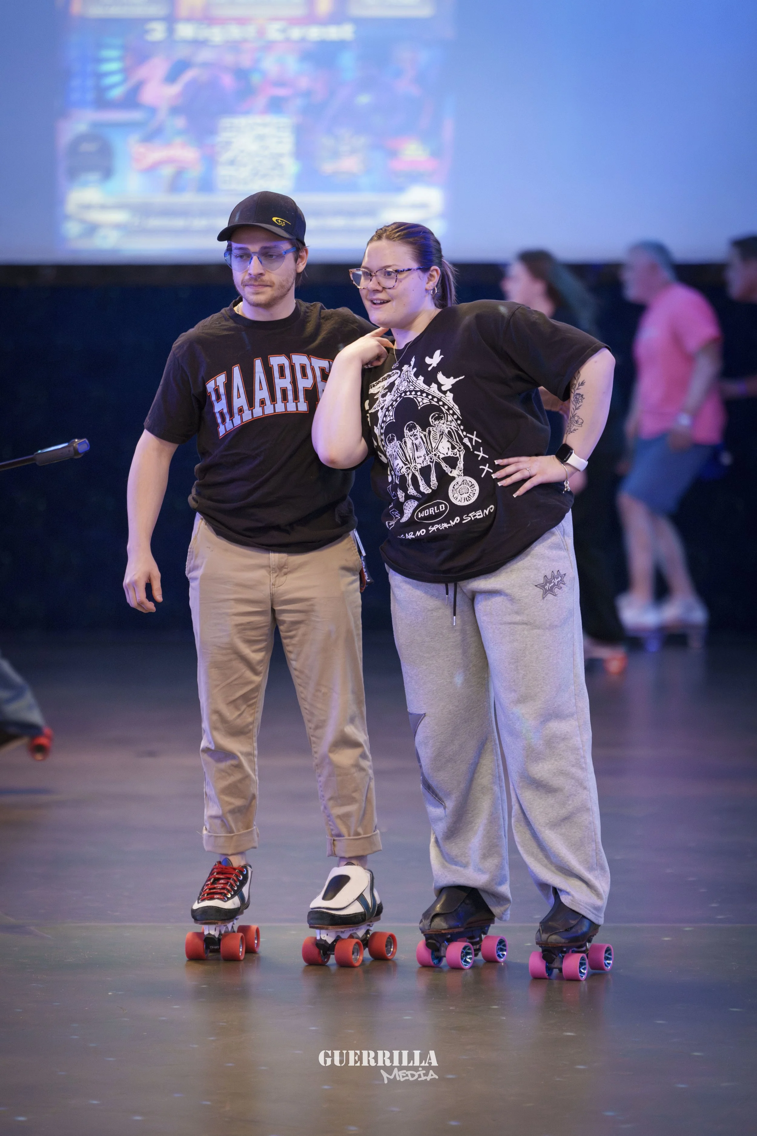 Two young adults roller skating indoors, standing close together, one with hand on hip, smiling and chatting, with a group of people blurred in the background.