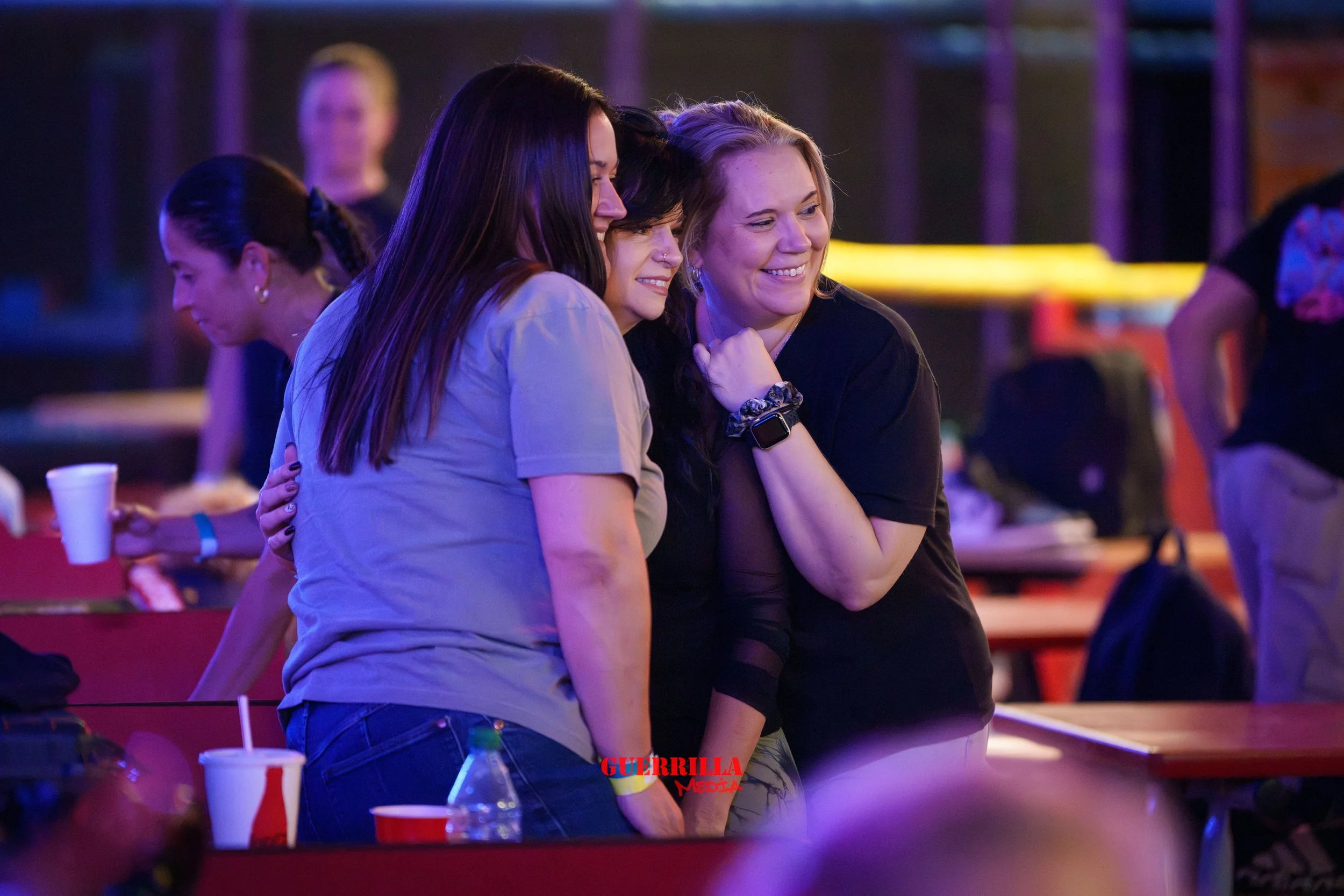 Three women smiling and embracing each other at an indoor event with colorful lighting, with other people and tables in the background.